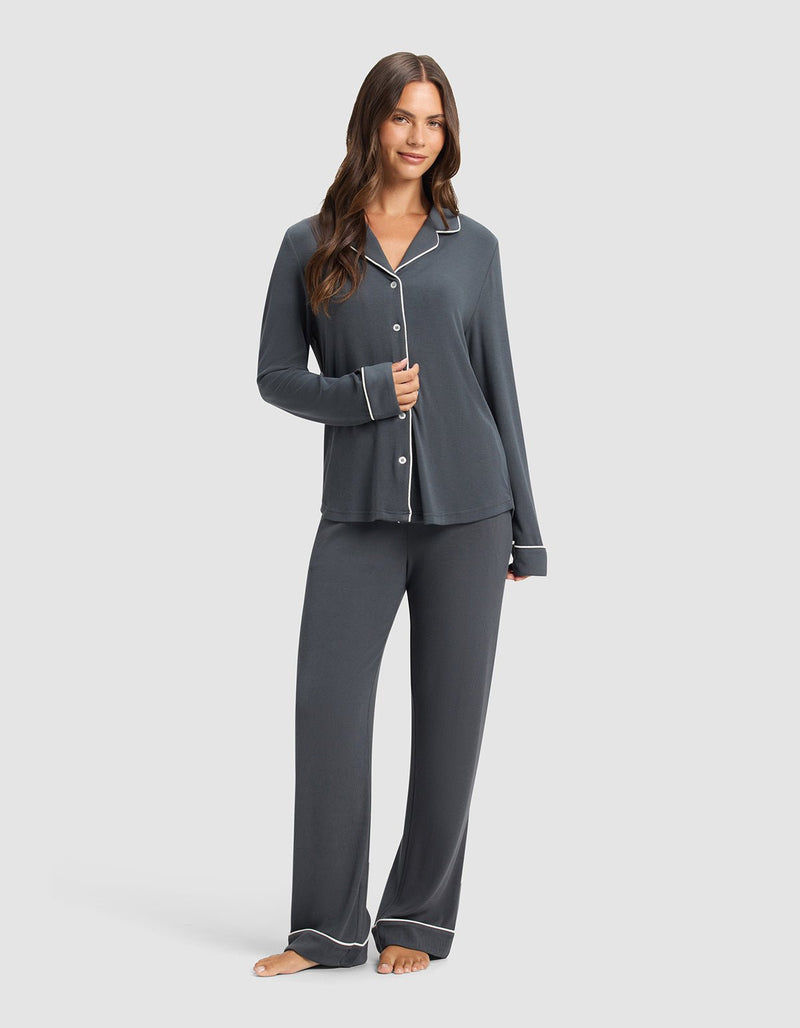 A woman with long, wavy hair and a relaxed expression stands barefoot, wearing Cozy Earth's Women's Bamboo Rib-Knit Classic Pajama Pant in dark gray with white piping, paired with a matching long-sleeve button-up top.