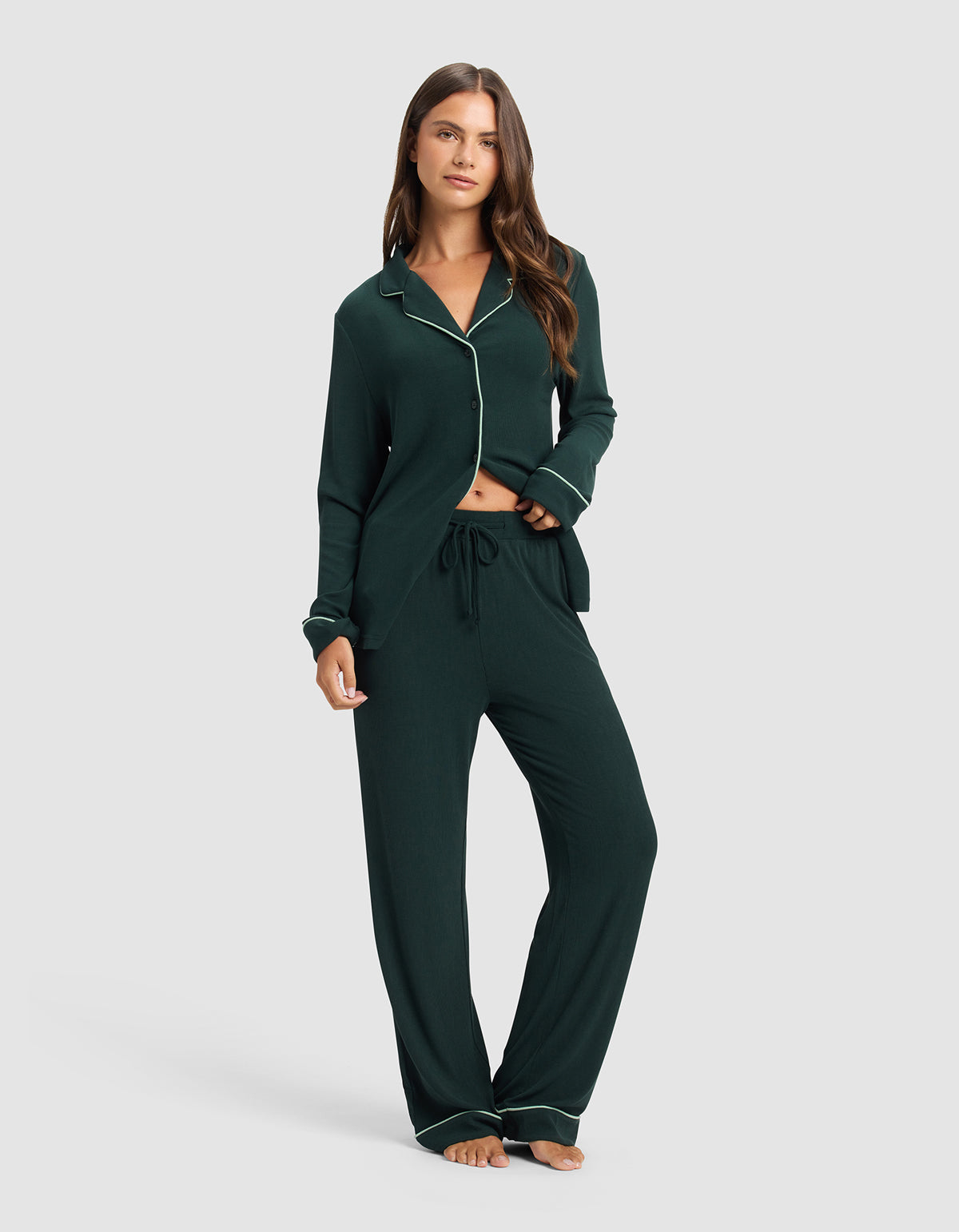 A woman stands barefoot in Cozy Earth's Women's Bamboo Rib-Knit Classic Pajama Pant, dark green with white piping and a drawstring waist. The long-sleeved top is buttoned and slightly open at the bottom. Background: plain light gray.|Color:Spruce
