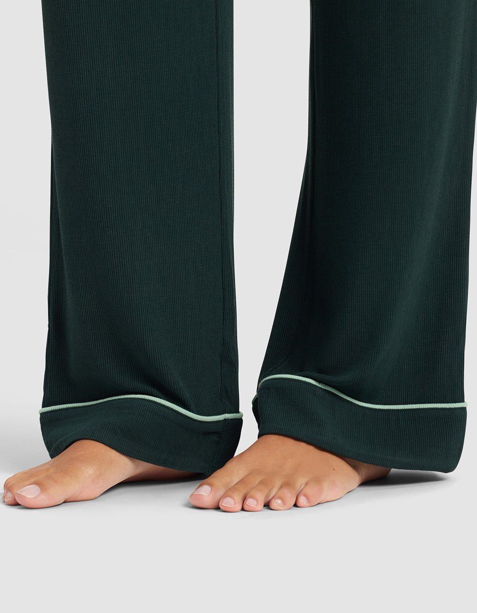 A close-up of bare feet wearing Cozy Earth's Women's Bamboo Rib-Knit Classic Pajama Pant in dark green with light green piping along the cuffs, standing on a light gray surface.