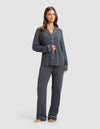 A woman stands barefoot in the Cozy Earth Women's Bamboo Rib-Knit Classic Long Sleeve Pajama Set, a dark gray matching set with white piping, featuring a button-up top and relaxed pants. She smiles slightly against a plain light background.|Color:Graphite
