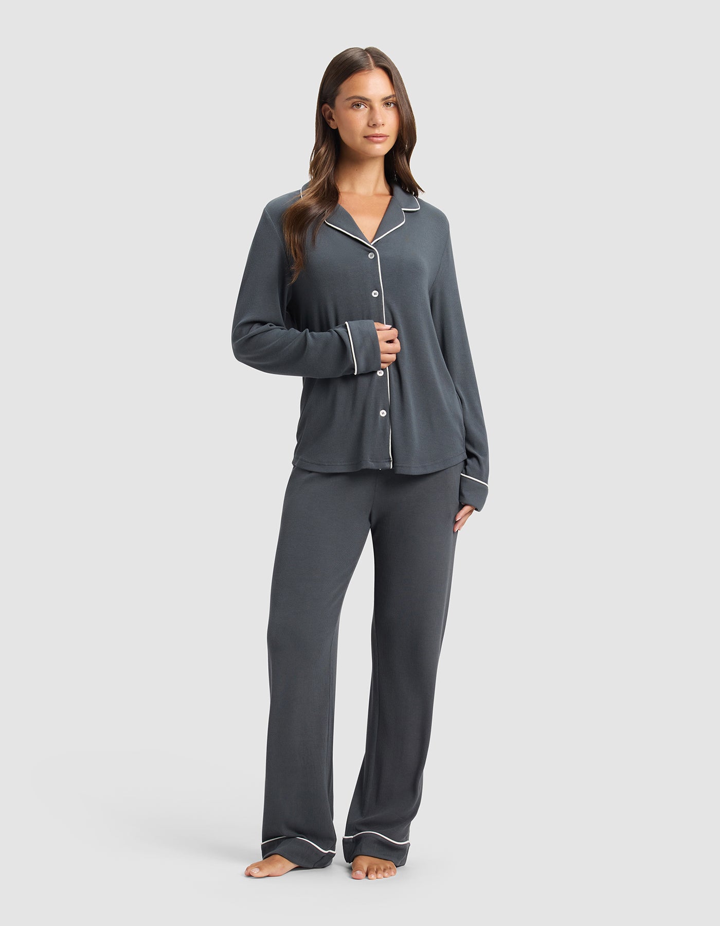 A woman stands barefoot in the Cozy Earth Women's Bamboo Rib-Knit Classic Long Sleeve Pajama Set, a dark gray matching set with white piping, featuring a button-up top and relaxed pants. She smiles slightly against a plain light background.|Color:Graphite