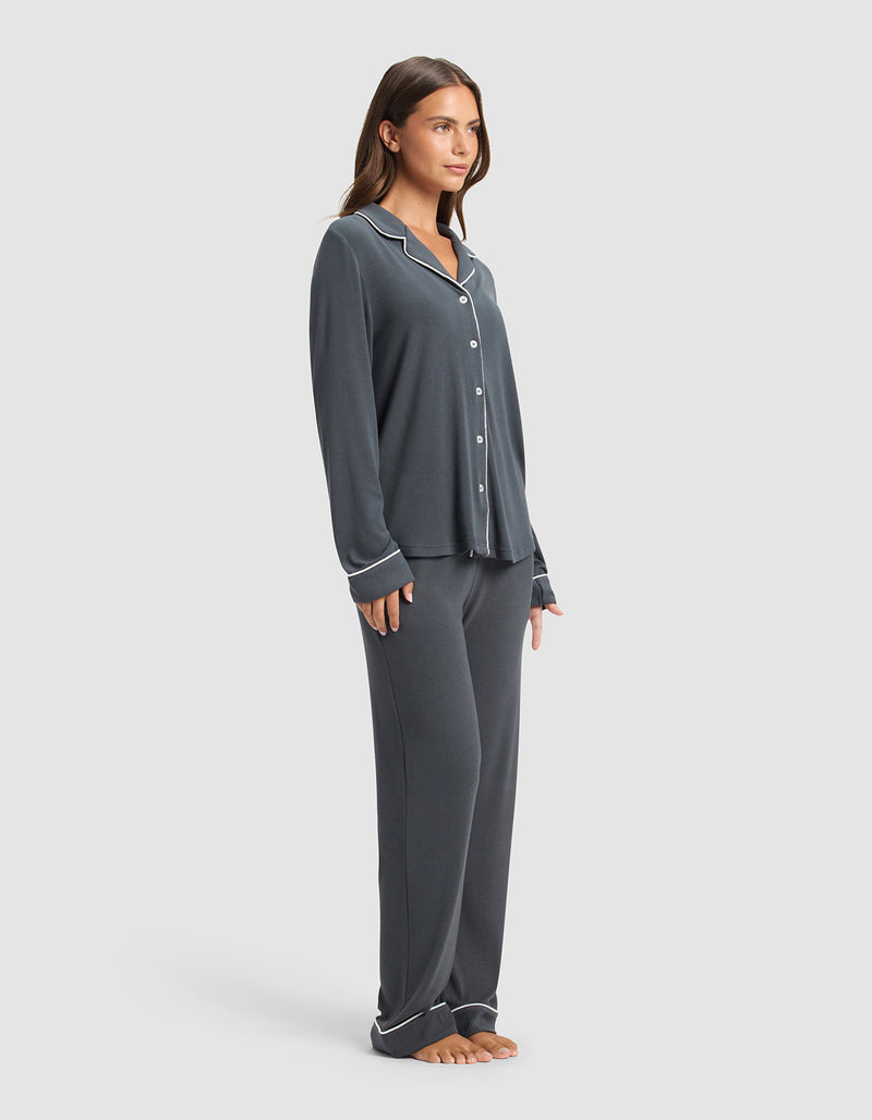 A woman stands barefoot against a plain light background, wearing Cozy Earth's Women's Bamboo Rib-Knit Classic Long Sleeve Pajama Set in dark gray with white trim, featuring a button-up top and loose pants.