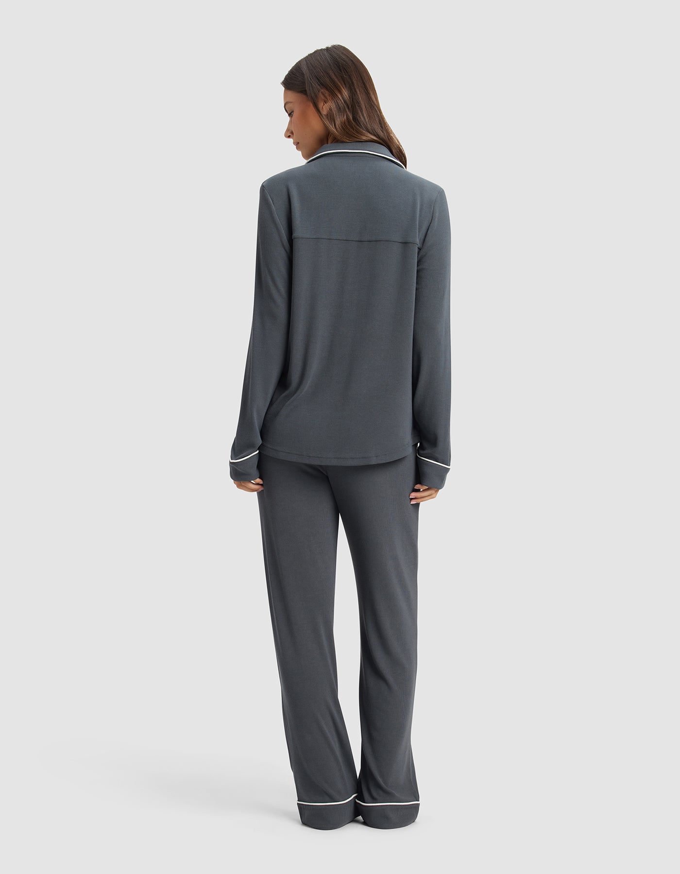 A woman faces away, wearing the Cozy Earth Women's Bamboo Rib-Knit Classic Long Sleeve Pajama Set in dark gray with white piping on the cuffs and pant hems, set against a plain light background.|Color:Graphite