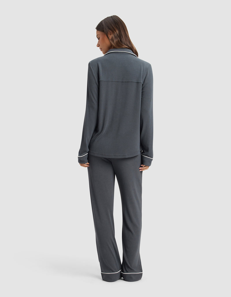 A woman faces away, wearing the Cozy Earth Women's Bamboo Rib-Knit Classic Long Sleeve Pajama Set in dark gray with white piping on the cuffs and pant hems, set against a plain light background.