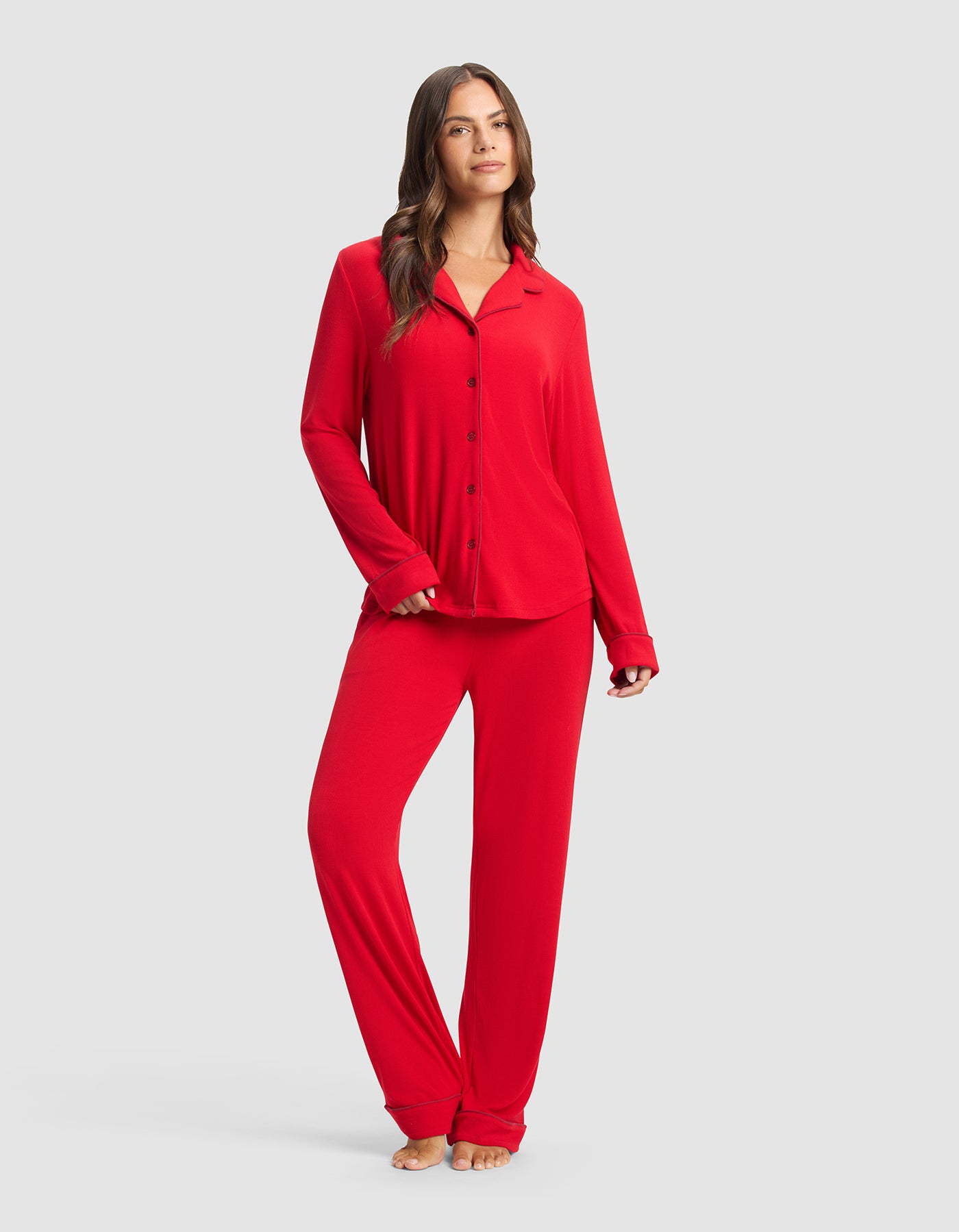 A woman with long brown hair stands barefoot against a light gray background, wearing Cozy Earth's Women's Bamboo Rib-Knit Classic Long Sleeve Pajama Set in red, featuring a button-down top and matching pants. She smiles gently.|Color:Crimson