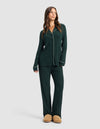A woman in the Cozy Earth Women's Bamboo Rib-Knit Classic Long Sleeve Pajama Set in dark green with white piping and tan slippers stands against a light gray background, gazing slightly aside with a relaxed expression.|Color:Spruce