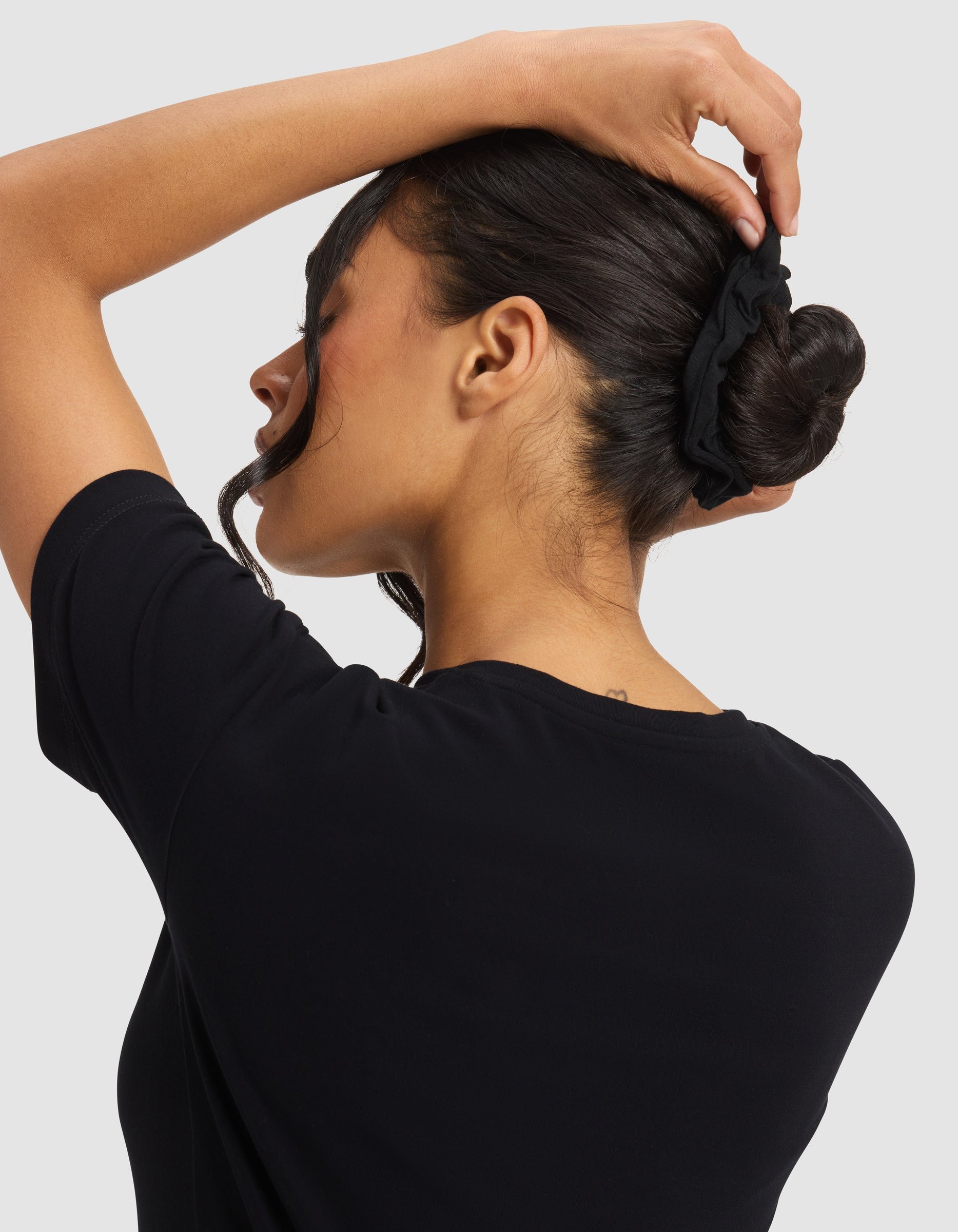 A woman with dark hair, wearing a black shirt, faces away and adjusts her bun with a Cozy Earth Bamboo Stretch-Knit Scrunchie. A loose strand falls by her face.