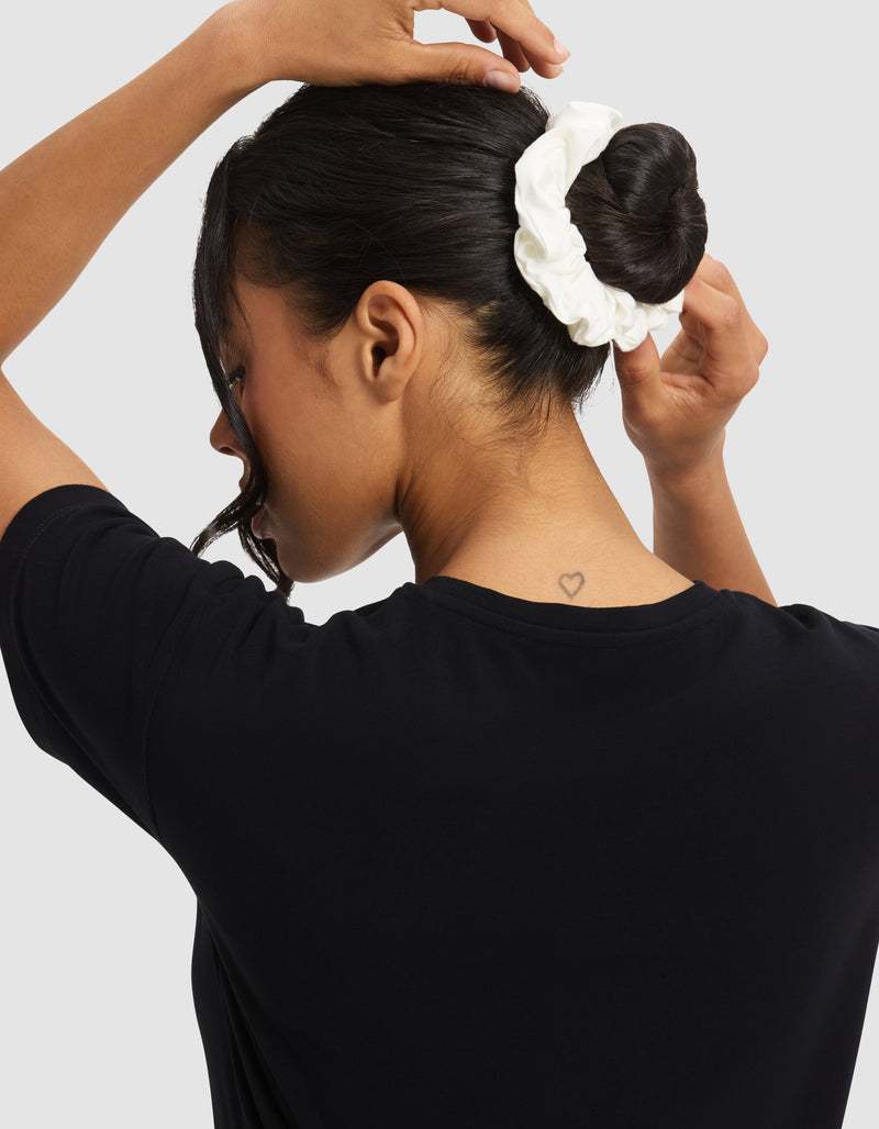 A person with dark hair ties their hair in a bun using a white Cozy Earth Bamboo Stretch-Knit Scrunchie. They wear a black shirt, have a small heart tattoo on the back of their neck, and stand against a light gray background.