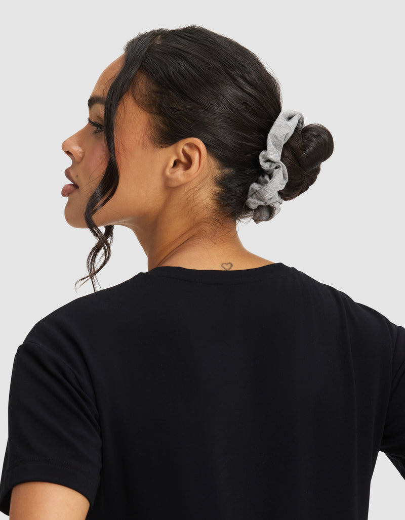A woman with dark hair in a bun secured by a Cozy Earth Bamboo Stretch-Knit Scrunchie, wearing a black shirt, is shown from the back and side. A small heart tattoo is visible on the nape of her neck.