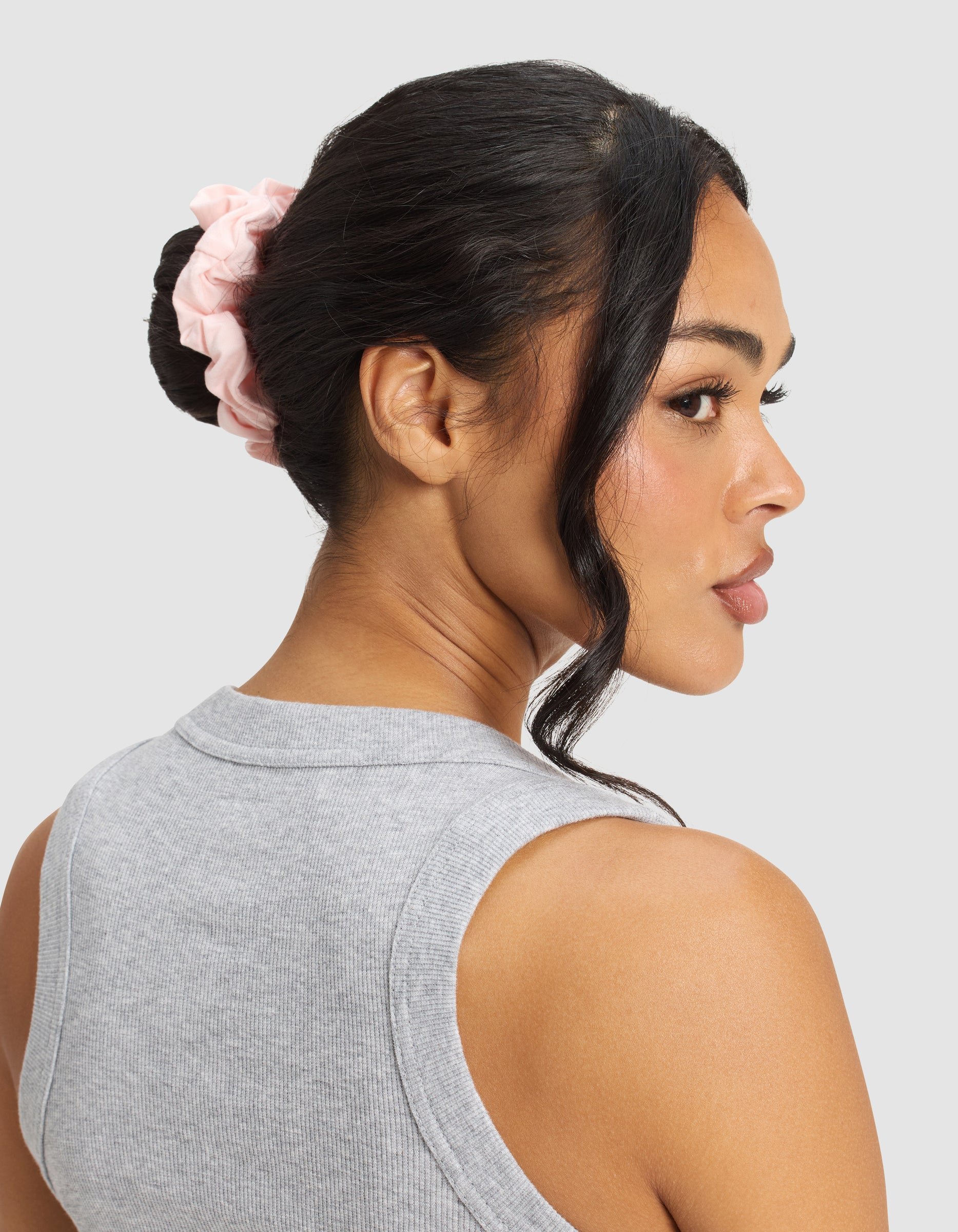 A woman with dark hair in a bun secured by a Cozy Earth Bamboo Stretch-Knit Scrunchie in light pink, wearing a sleeveless gray top, looks over her shoulder against a light gray background.
