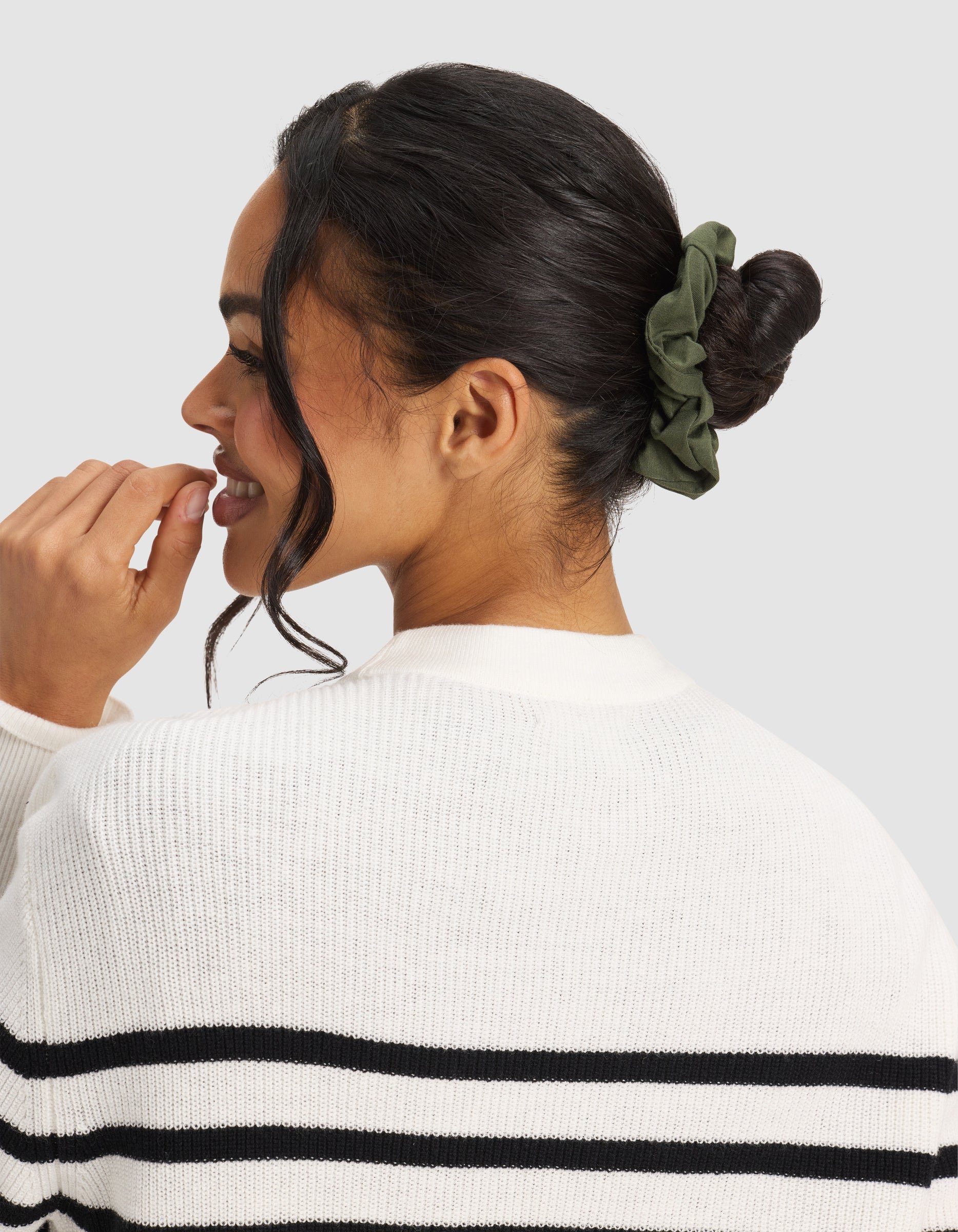 A woman with dark hair in a bun, secured by a Cozy Earth Bamboo Stretch-Knit Scrunchie in olive green, wears a white sweater with black stripes and smiles to the side against a plain background.