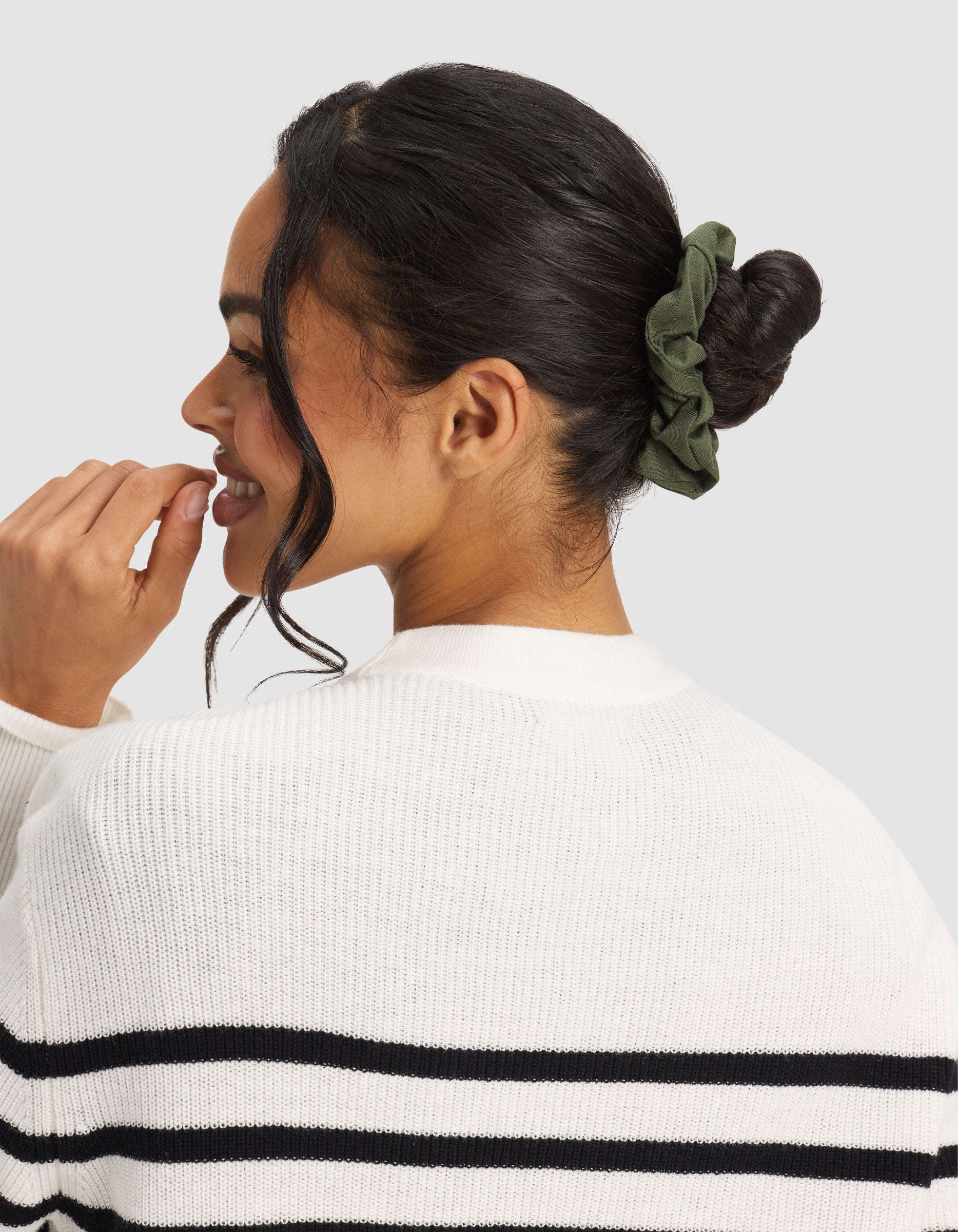 A woman with dark hair in a bun, secured by a Cozy Earth Bamboo Stretch-Knit Scrunchie in olive green, wears a white sweater with black stripes and smiles to the side against a plain background.