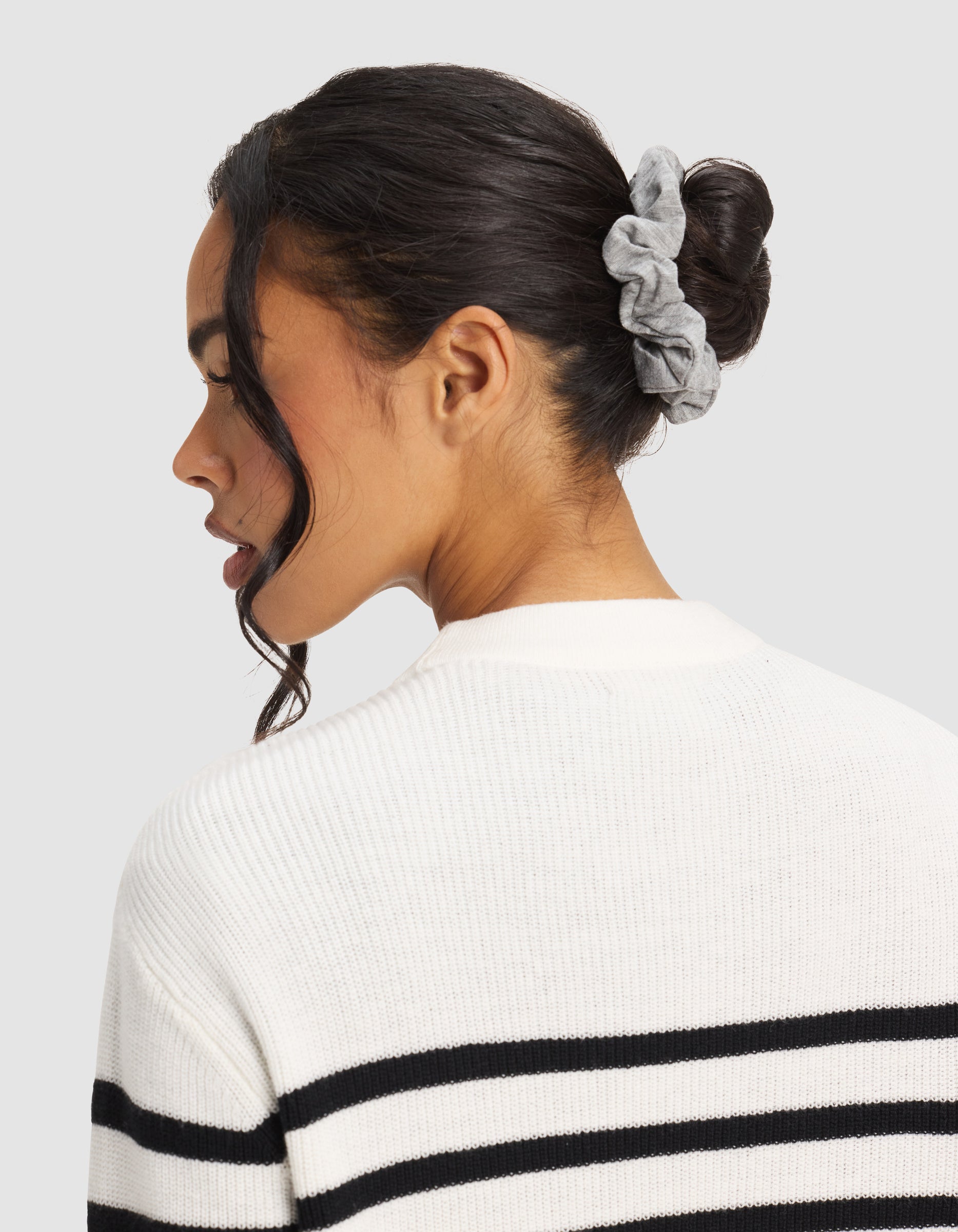 A woman with dark hair in a bun, secured by a Cozy Earth Bamboo Stretch-Knit Scrunchie, wears a white sweater with black stripes and faces away against a plain background.