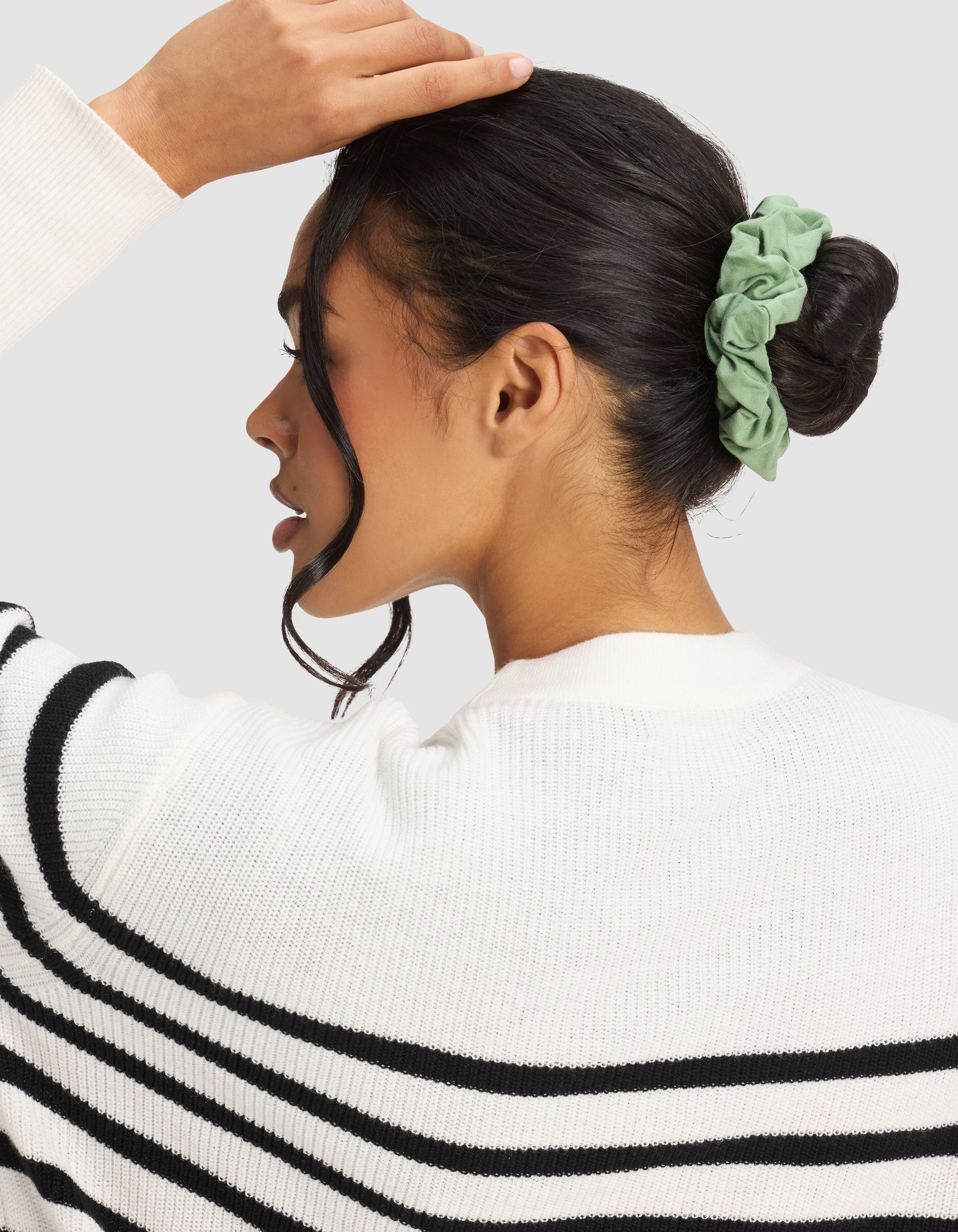 A woman with dark hair in a bun, secured by a Cozy Earth Bamboo Stretch-Knit Scrunchie in green, wears a white sweater with black stripes and is shown from the side against a light gray background.