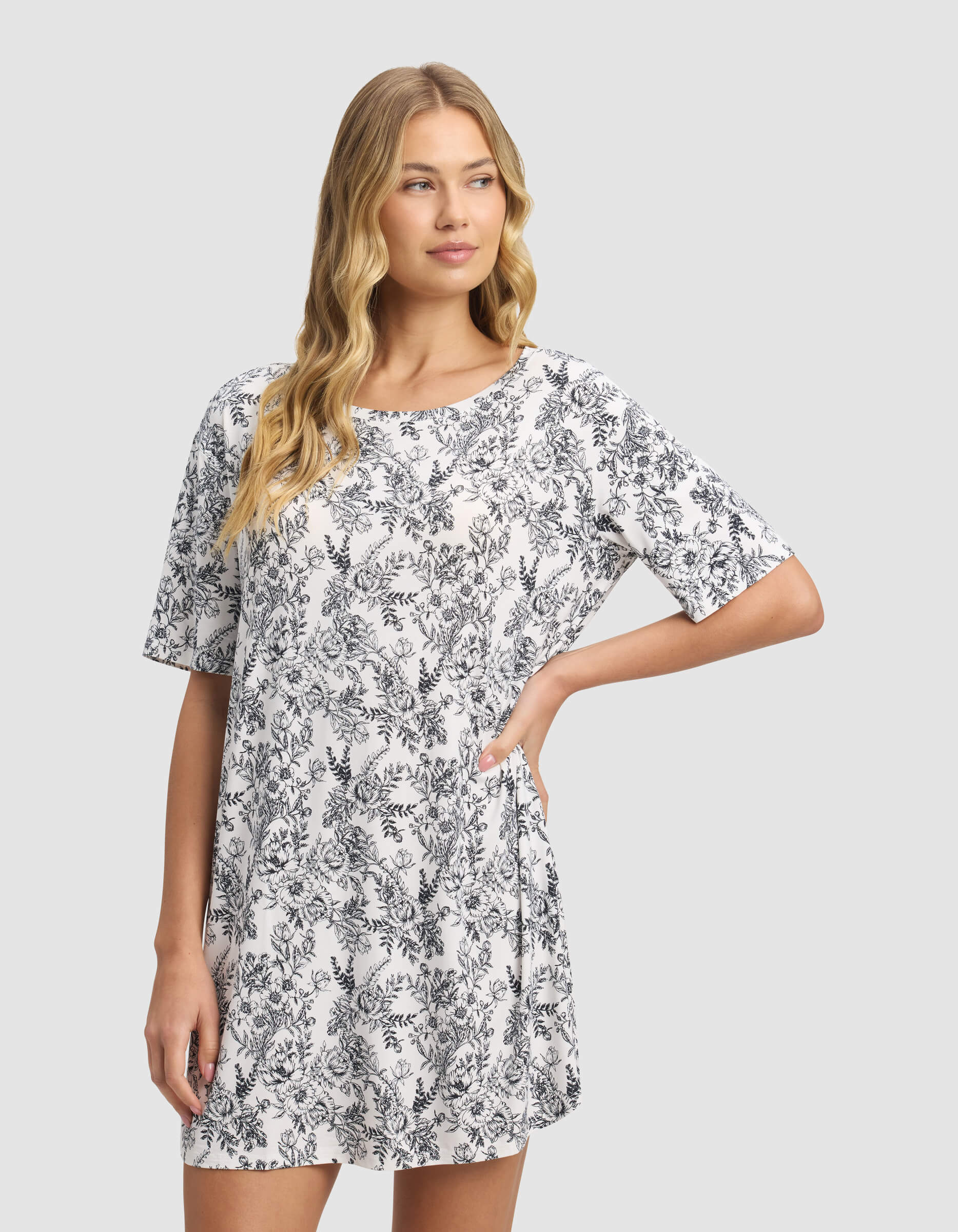 A woman with long blonde hair models the Cozy Earth Women's Bamboo Stretch-Knit Sleep Dress, featuring short sleeves and a white fabric with a black floral pattern. She stands against a plain light gray background, looking to the side.