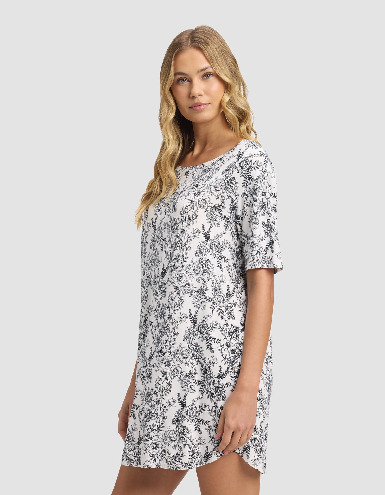 A woman with long blonde hair models the Cozy Earth Women's Bamboo Stretch-Knit Sleep Dress, featuring short sleeves and a dark floral pattern, standing against a plain light background and gazing slightly to the side.
