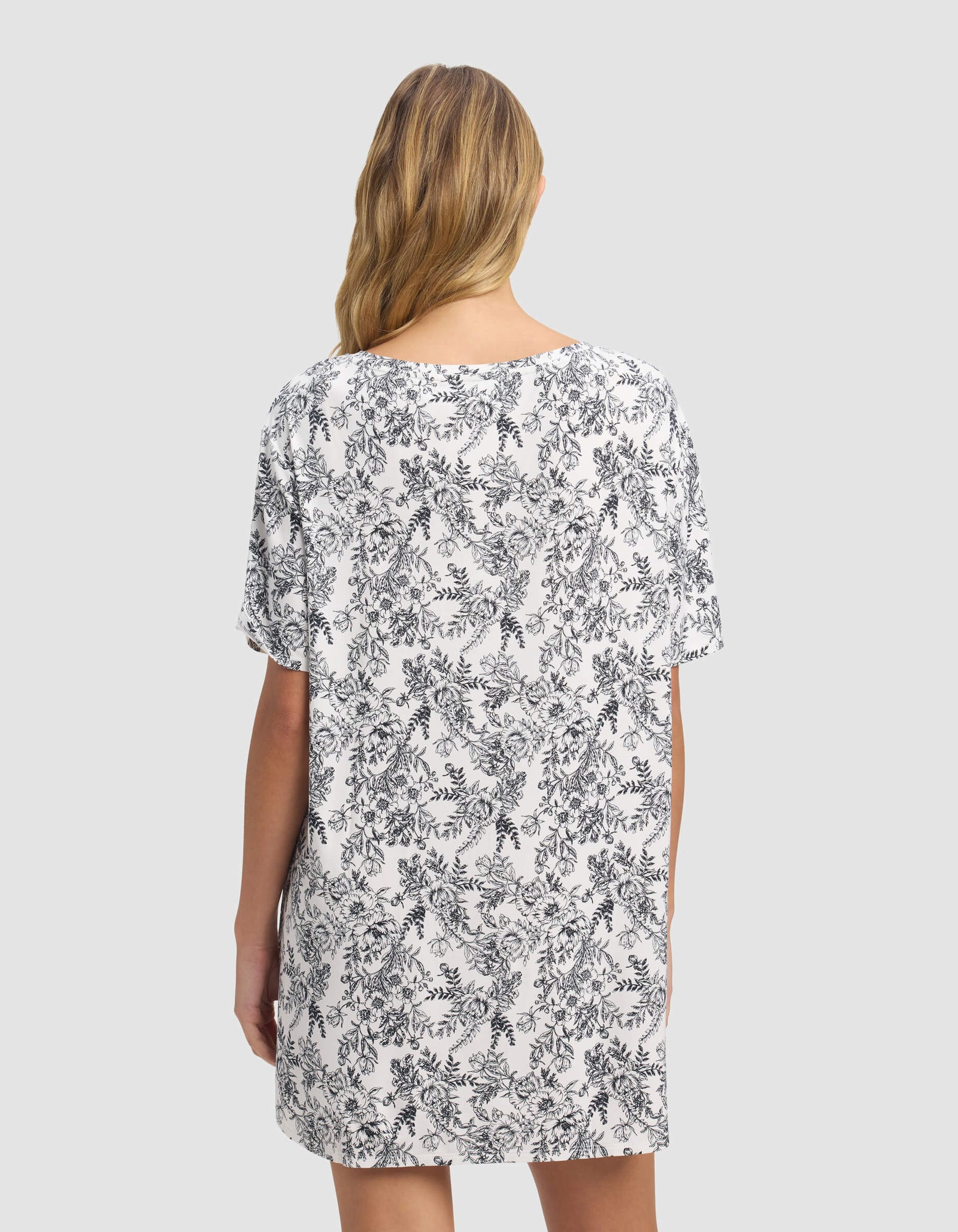 A woman with blonde hair stands facing away, wearing the Cozy Earth Women's Bamboo Stretch-Knit Sleep Dress in white with a black floral pattern on a light gray background.