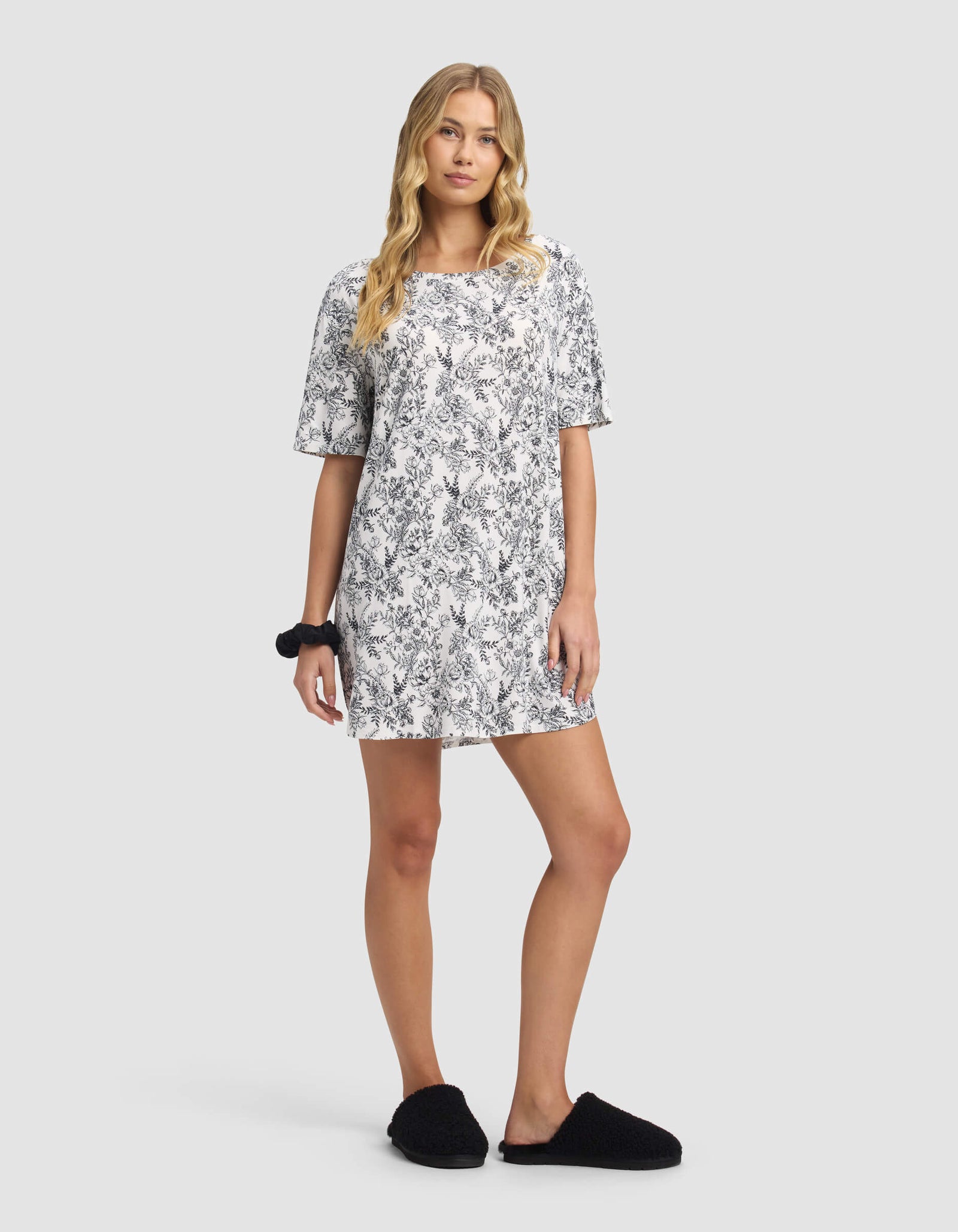 A woman with long, wavy blonde hair wears the Cozy Earth Women's Bamboo Stretch-Knit Sleep Dress in white with a black floral pattern, paired with black slippers and a scrunchie, standing against a plain light background.