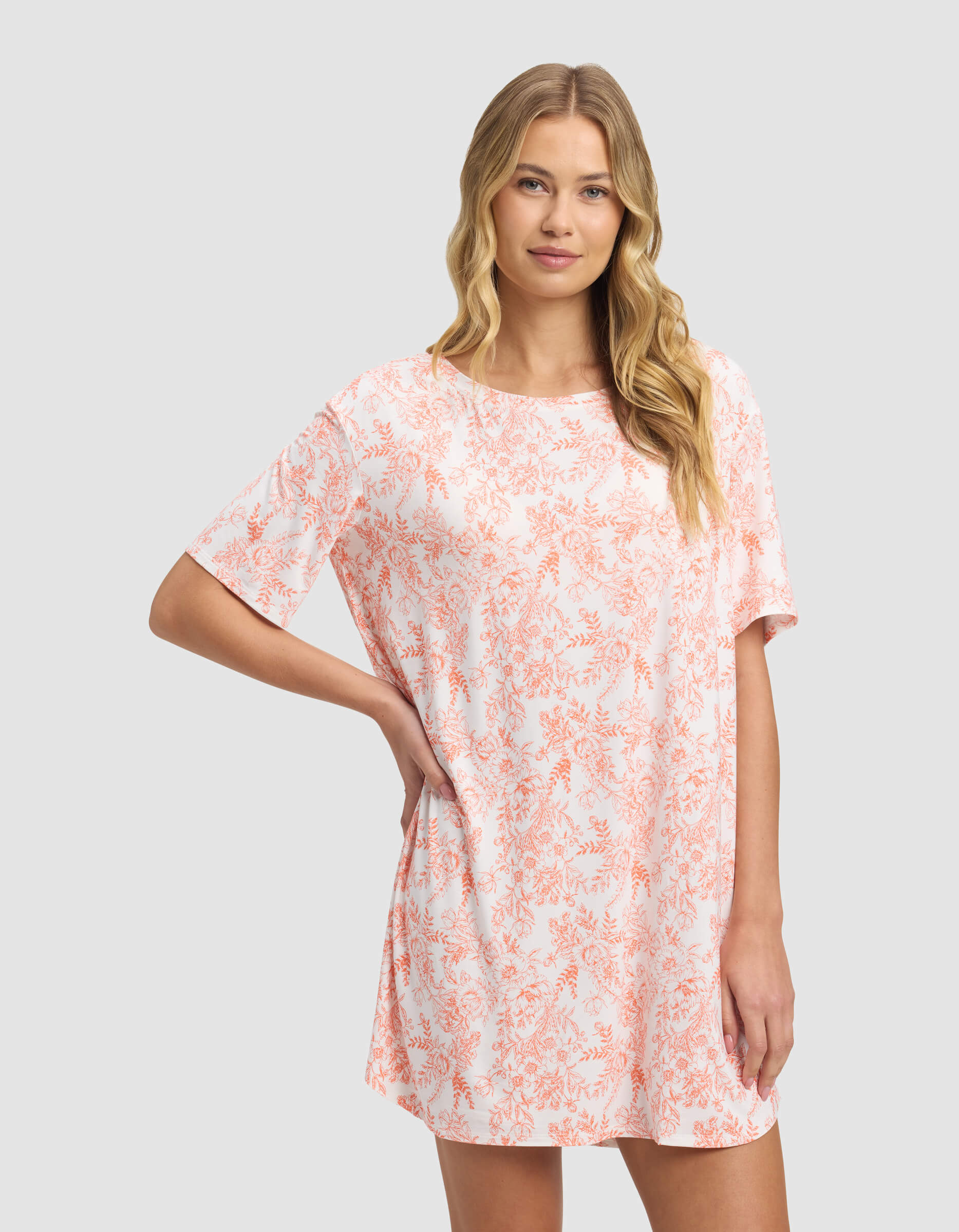 A woman wearing the Cozy Earth Women's Bamboo Stretch-Knit Sleep Dress stands against a light gray background, gently smiling with one hand on her hip.|Color:Melon Toile