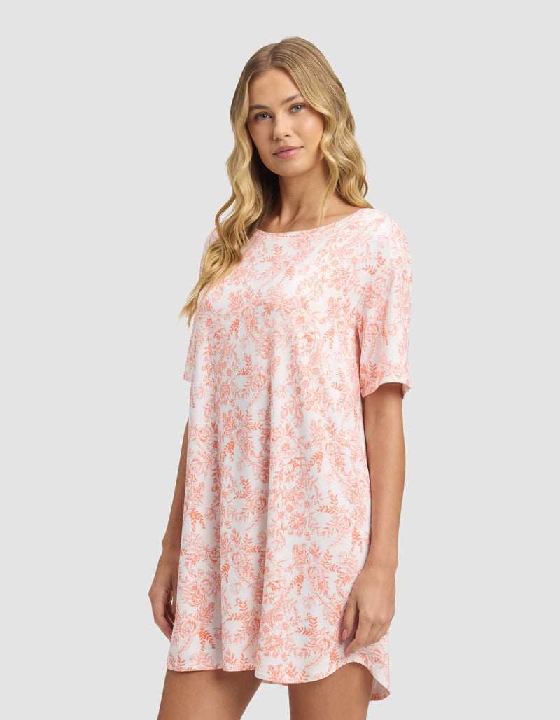 A woman with long blonde hair models the Cozy Earth Women's Bamboo Stretch-Knit Sleep Dress, a loose, short-sleeved light pink dress with an orange floral pattern, against a plain light gray background.