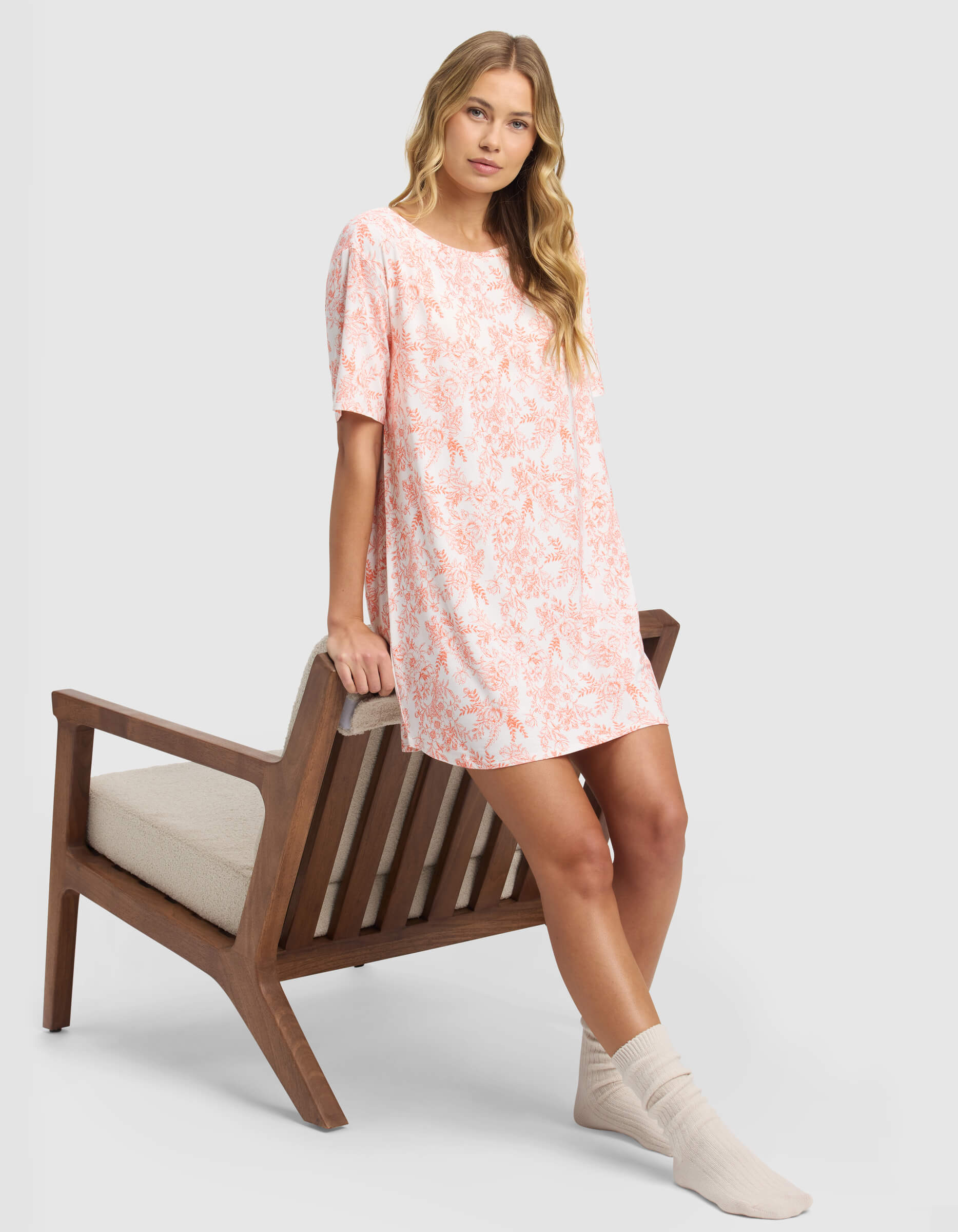 A woman in the Cozy Earth Women's Bamboo Stretch-Knit Sleep Dress, featuring loose short sleeves and a pink floral print, with cream socks, leans against a wooden chair with a neutral cushion against a light gray background.|Color:Melon Toile
