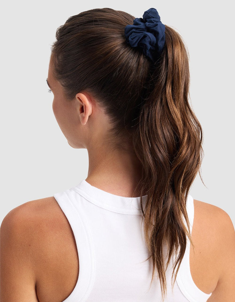 A woman with long, wavy brown hair in a ponytail, styled with a Cozy Earth Bamboo Stretch-Knit Scrunchie in navy blue, wears a white sleeveless top and faces away against a plain light gray background.