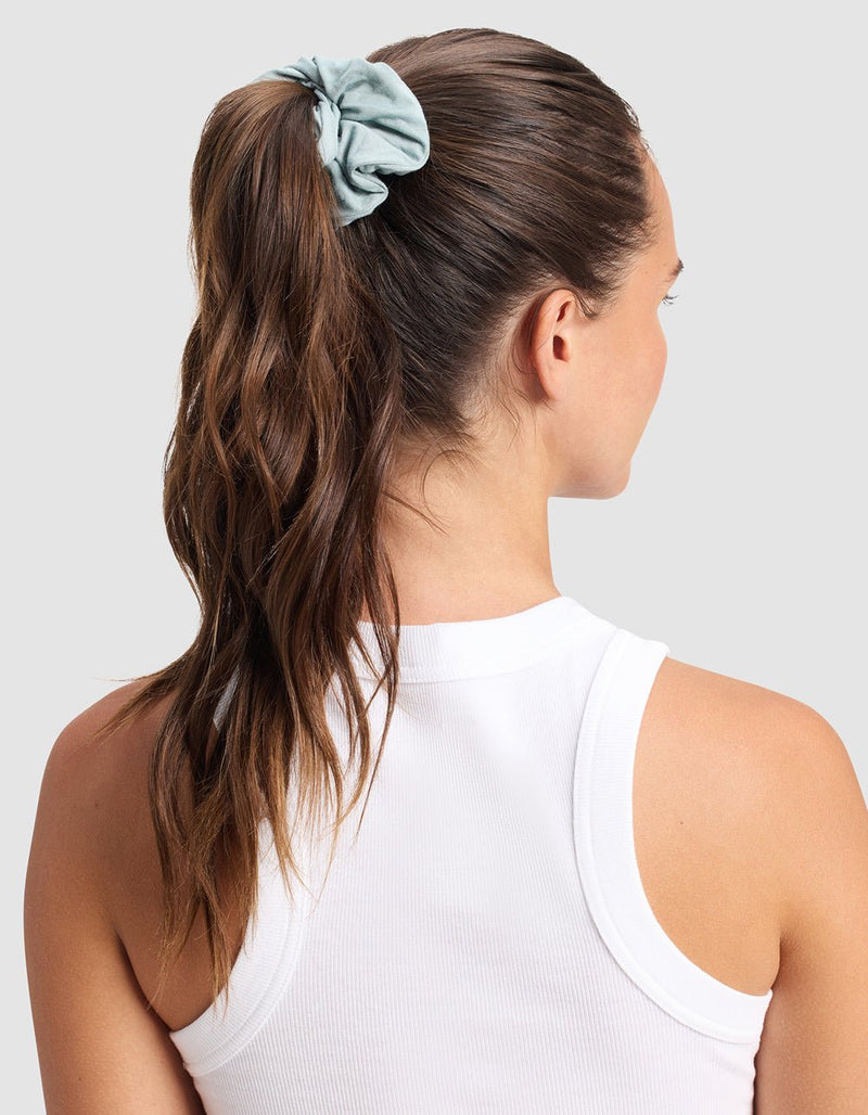A woman with long brown hair in a high ponytail, secured by Cozy Earth’s Bamboo Stretch-Knit Scrunchie in light blue, wears a white sleeveless top and faces away against a plain light grey background.