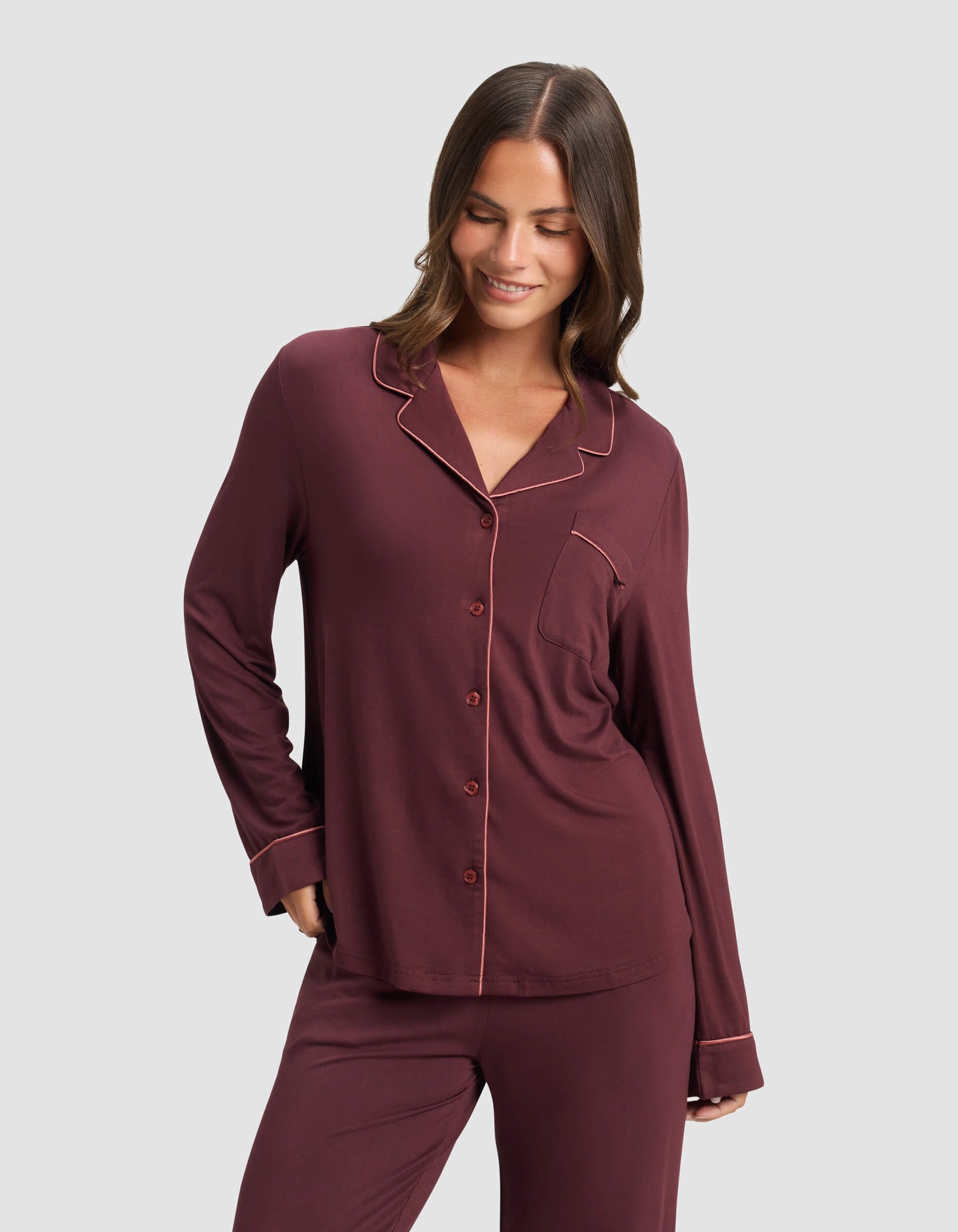 A woman wears the Cozy Earth Women's Bamboo Stretch-Knit Long Sleeve Pajama Top in burgundy, featuring light piping, standing against a plain light gray background and smiling while looking down.