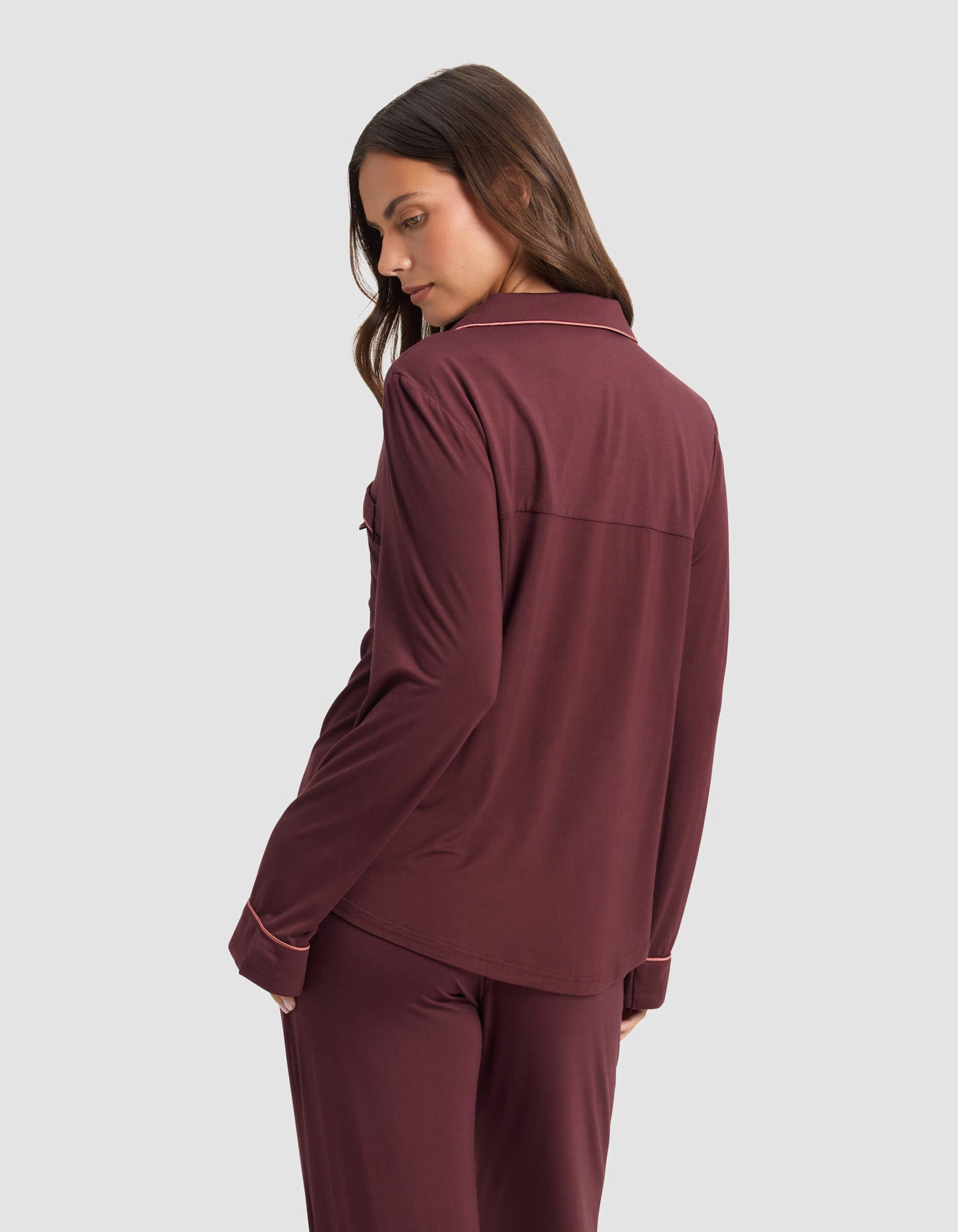 A woman with loose hair stands sideways, looking over her shoulder, wearing the Cozy Earth Women's Bamboo Stretch-Knit Long Sleeve Pajama Top in burgundy against a plain light gray background.
