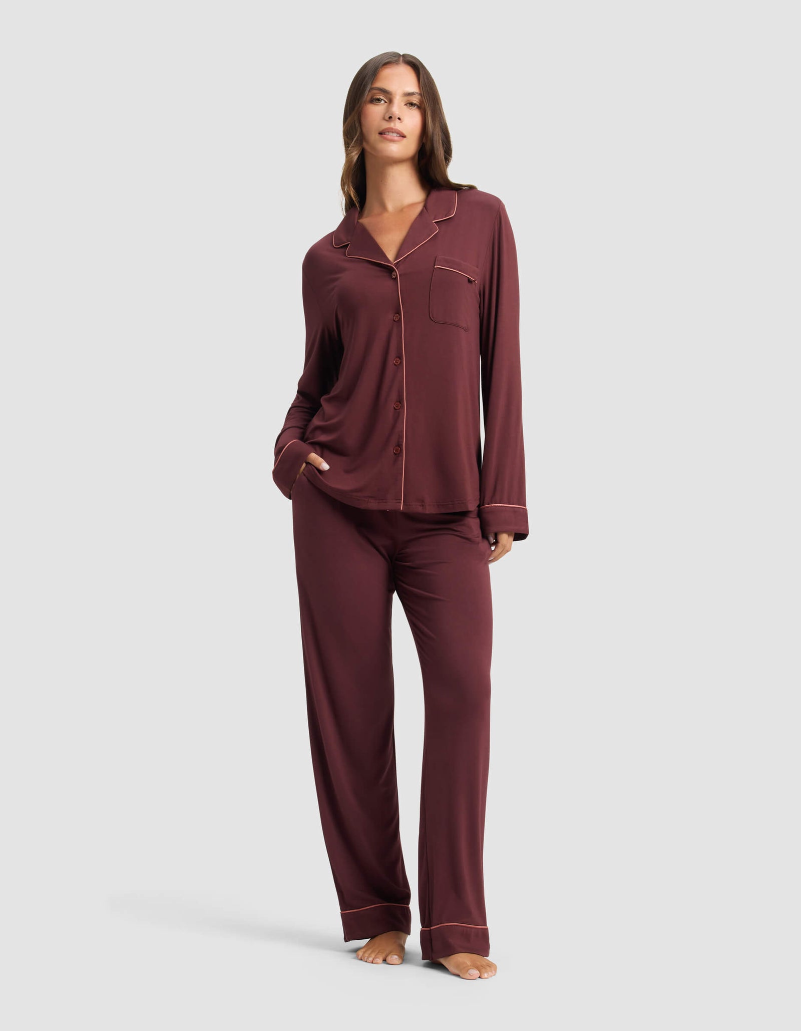 A woman stands barefoot against a light background, wearing the Cozy Earth Women's Bamboo Stretch-Knit Long Sleeve Pajama Top in maroon, featuring buttons and a chest pocket, paired with matching pants and posing with one hand in her pocket.