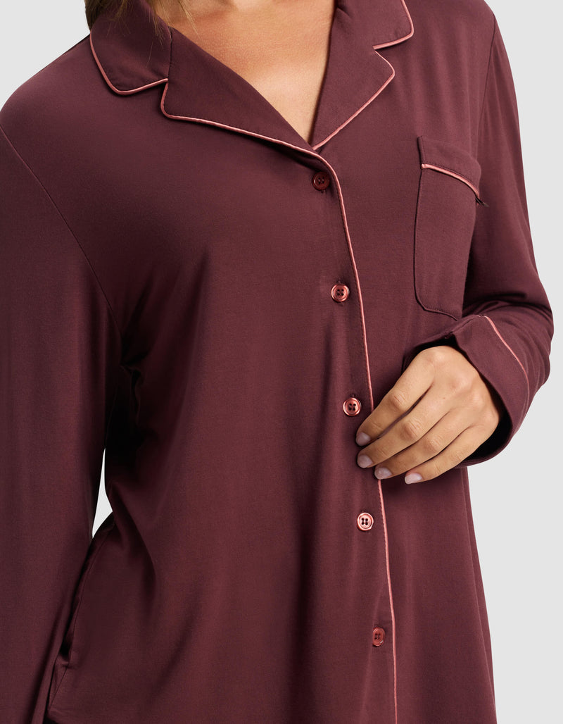 A woman wears the Cozy Earth Women's Bamboo Stretch-Knit Long Sleeve Pajama Top in maroon with pink piping, front buttons, and a chest pocket; her hand rests near the lower buttons.