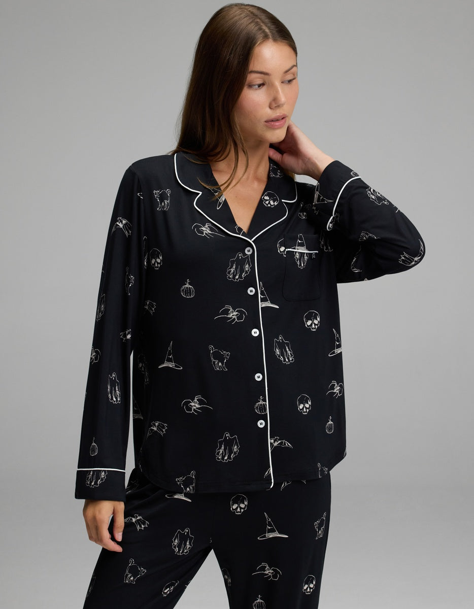 A woman wearing the Cozy Earth Women’s Bamboo Stretch-Knit Long Sleeve Pajama Set, featuring black fabric with white piping and outlined designs, stands against a plain grey background, gazing slightly to the side with one hand gently touching her neck.