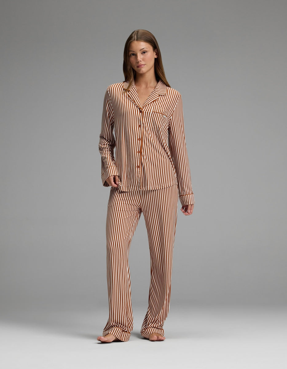 A woman stands barefoot against a plain gray background, wearing the Cozy Earth Women’s Bamboo Stretch-Knit Long Sleeve Pajama Top, styled with matching brown and white vertical striped pajama pants.