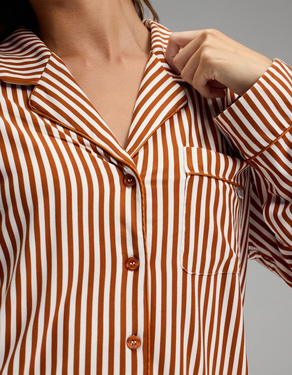 A person wearing the Cozy Earth Women’s Bamboo Stretch-Knit Long Sleeve Pajama Top in a brown and white vertical stripe pattern, featuring a collar and chest pocket, holds the collar with one hand. The face is not visible.