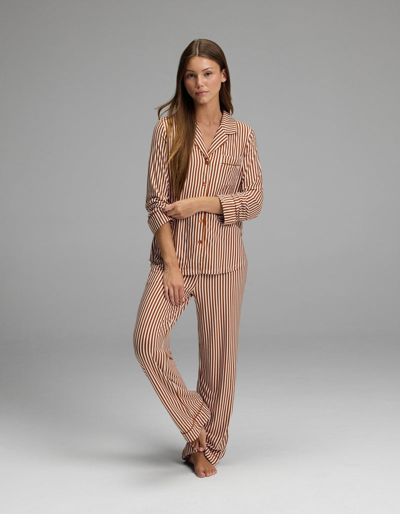 A woman stands barefoot on a gray background, wearing the Cozy Earth Women’s Bamboo Stretch-Knit Long Sleeve Pajama Top in beige and brown stripes, her arms loosely crossed and her expression relaxed.