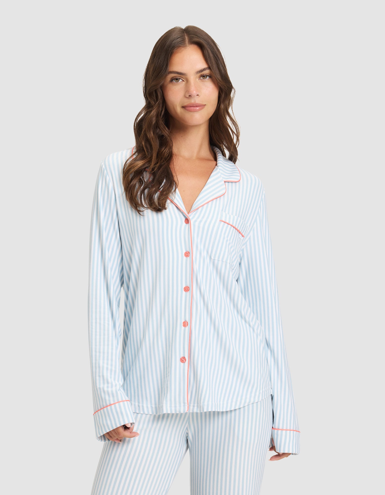 A woman with long brown hair models the Cozy Earth Women's Bamboo Stretch-Knit Long Sleeve Pajama Set, featuring light blue and white stripes with red buttons and trim, standing against a plain light gray background and smiling gently. 