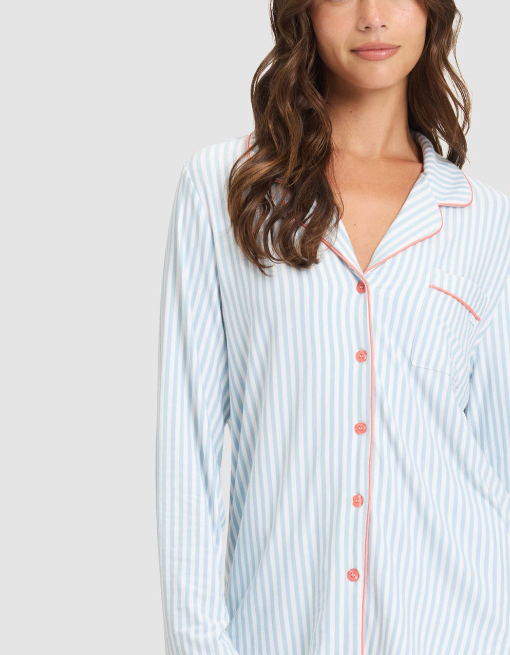 A woman with long brown hair models the Cozy Earth Women's Bamboo Stretch-Knit Long Sleeve Pajama Set, featuring light blue and white stripes with red buttons and trim, standing against a plain light gray background and smiling gently. |Color:Market Stripe in Lake