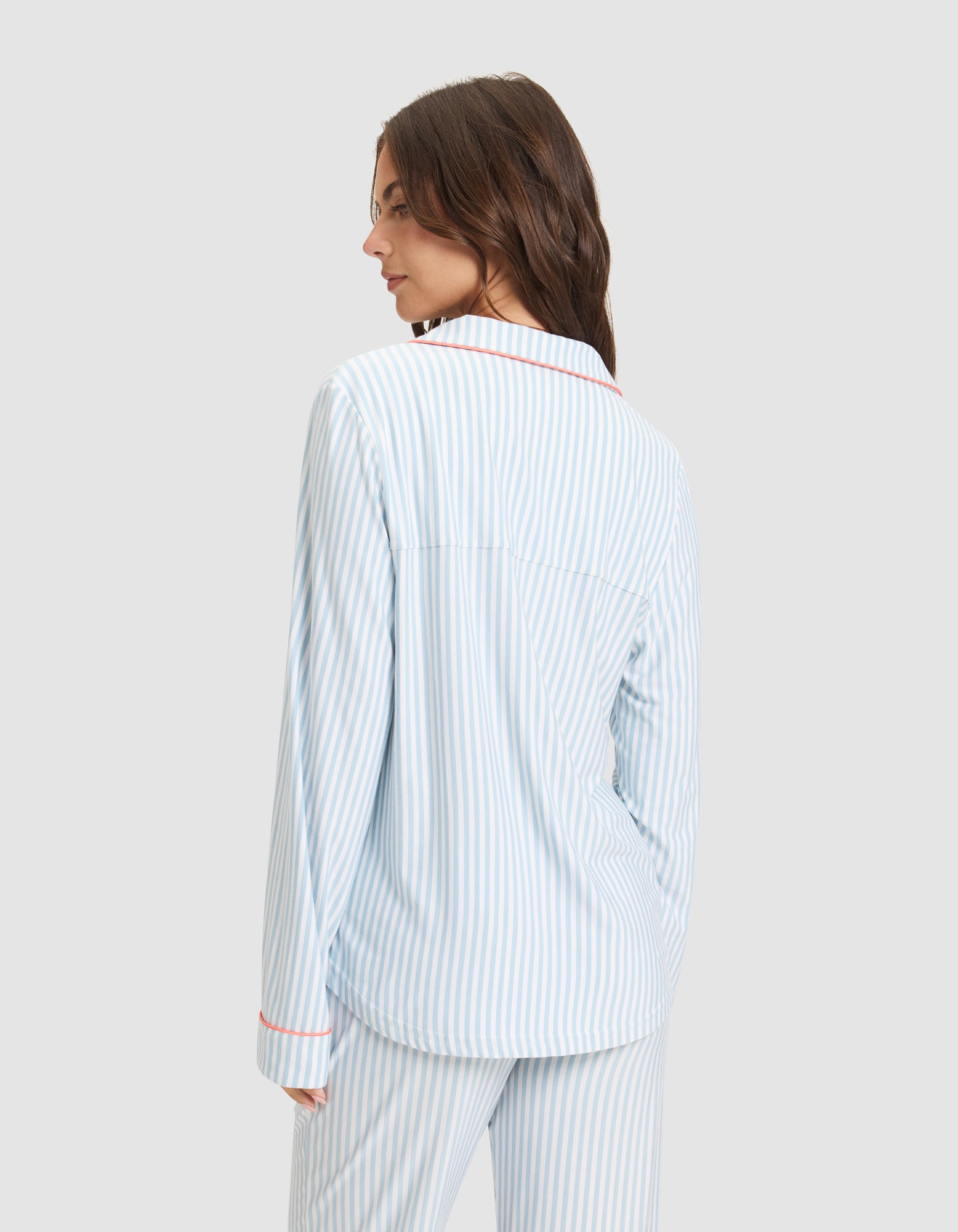 A woman with long brown hair is seen from behind wearing the Cozy Earth Women's Bamboo Stretch-Knit Long Sleeve Pajama Set, featuring light blue and white stripes with red trim, against a plain light gray background. 