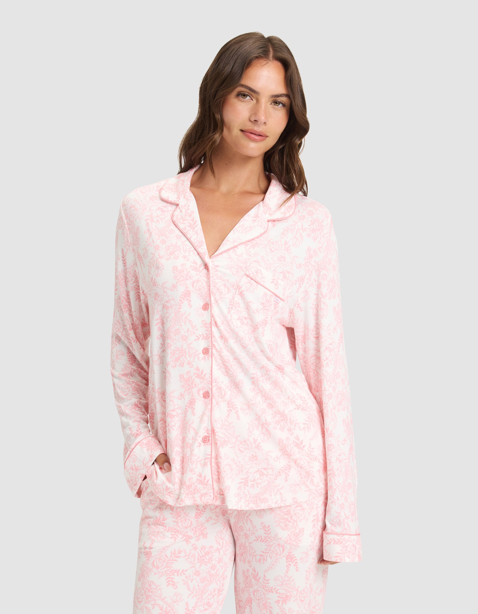 A woman models the Cozy Earth Women's Bamboo Stretch-Knit Long Sleeve Pajama Set in light pink and white, standing against a plain light gray background with one hand in her pocket, smiling softly at the camera. 