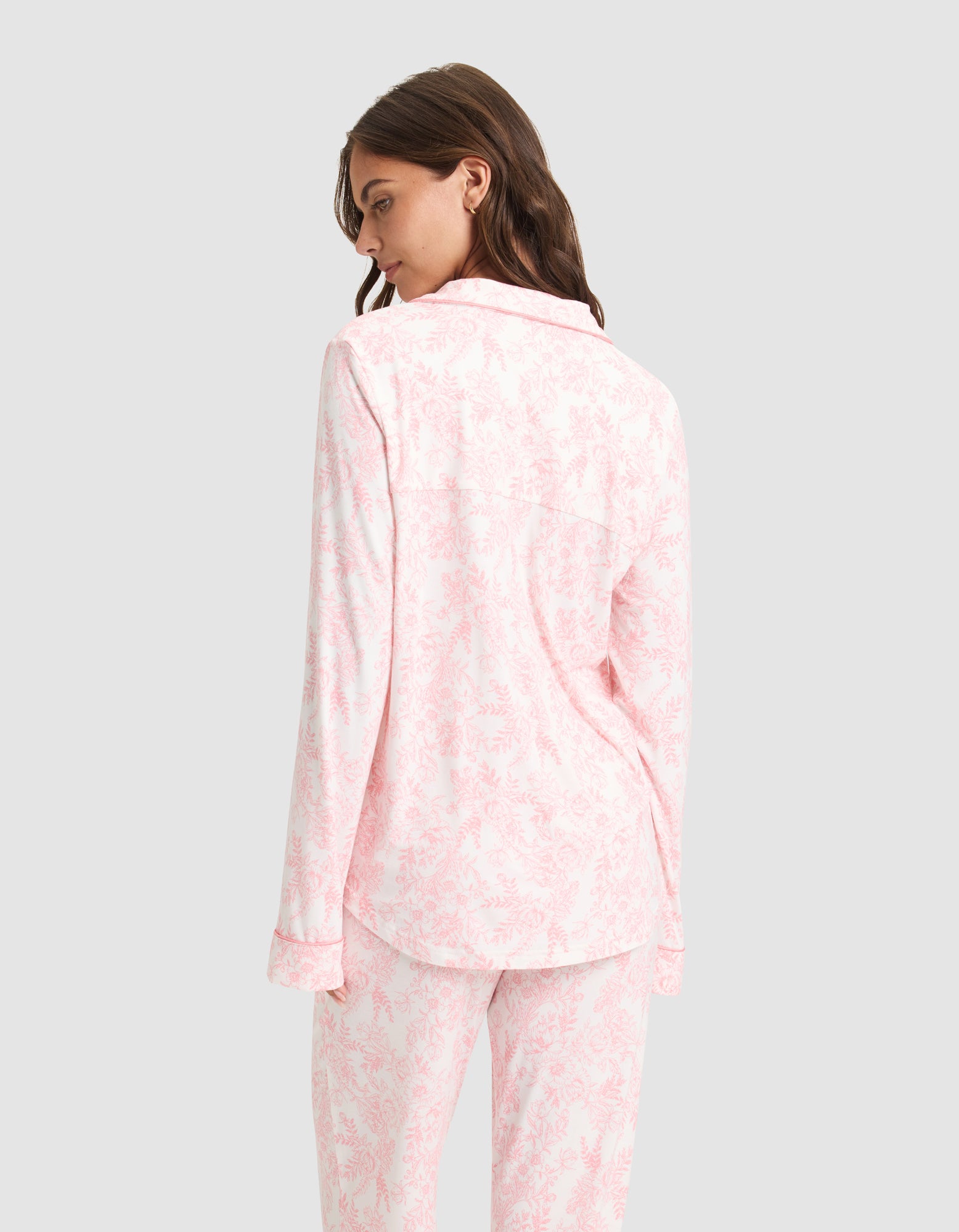 A woman models the Women's Bamboo Stretch-Knit Long Sleeve Pajama Set by Cozy Earth in light pink and white, featuring a collared, long-sleeve top and loose-fit pants. She is shown from the back against a plain light gray background. 