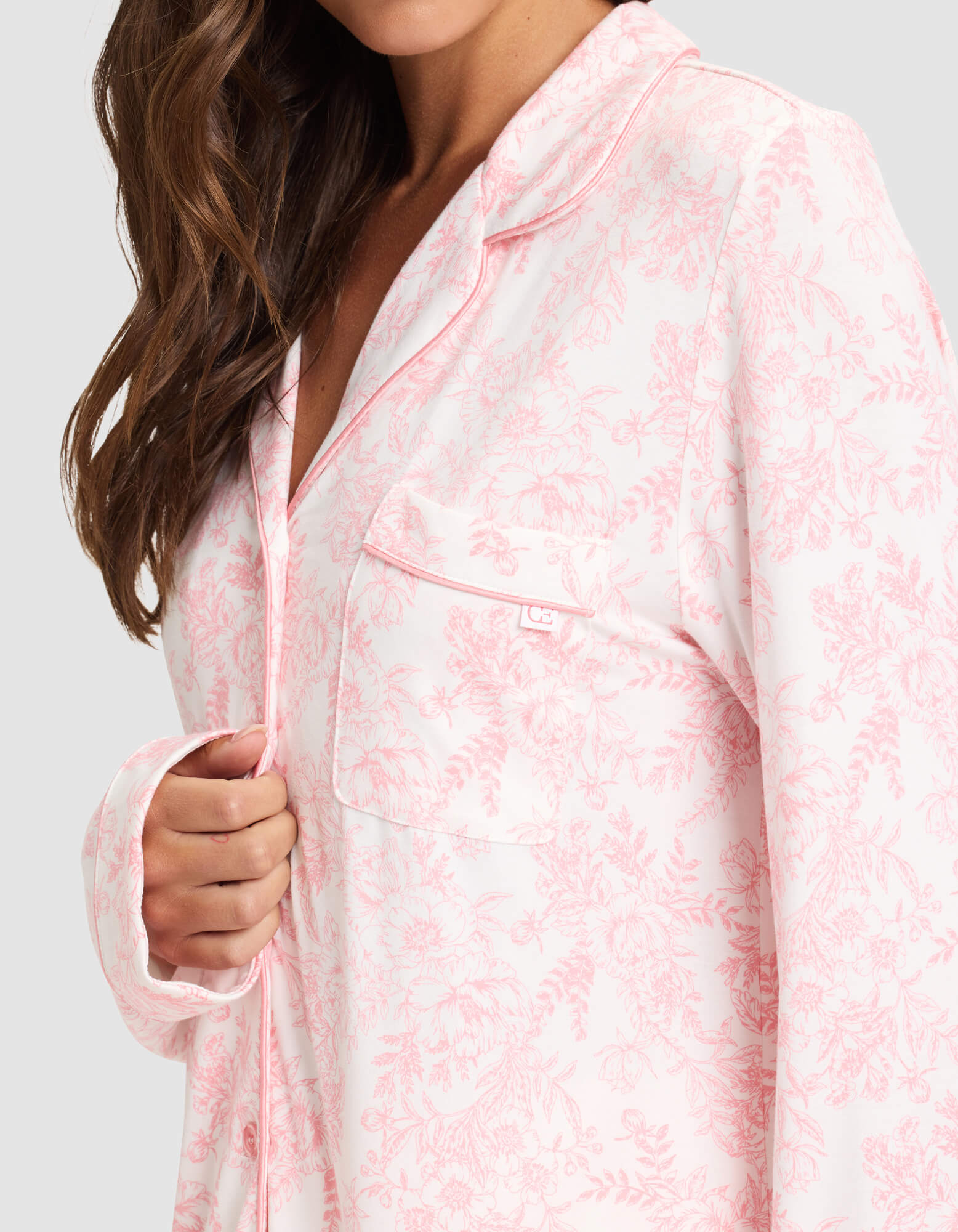 A woman with long brown hair wears the Cozy Earth Women's Bamboo Stretch-Knit Long Sleeve Pajama Set in a light pink and white floral pattern. She holds the top's edge, showing its chest pocket and piped trim. Her face is partially out of frame. 