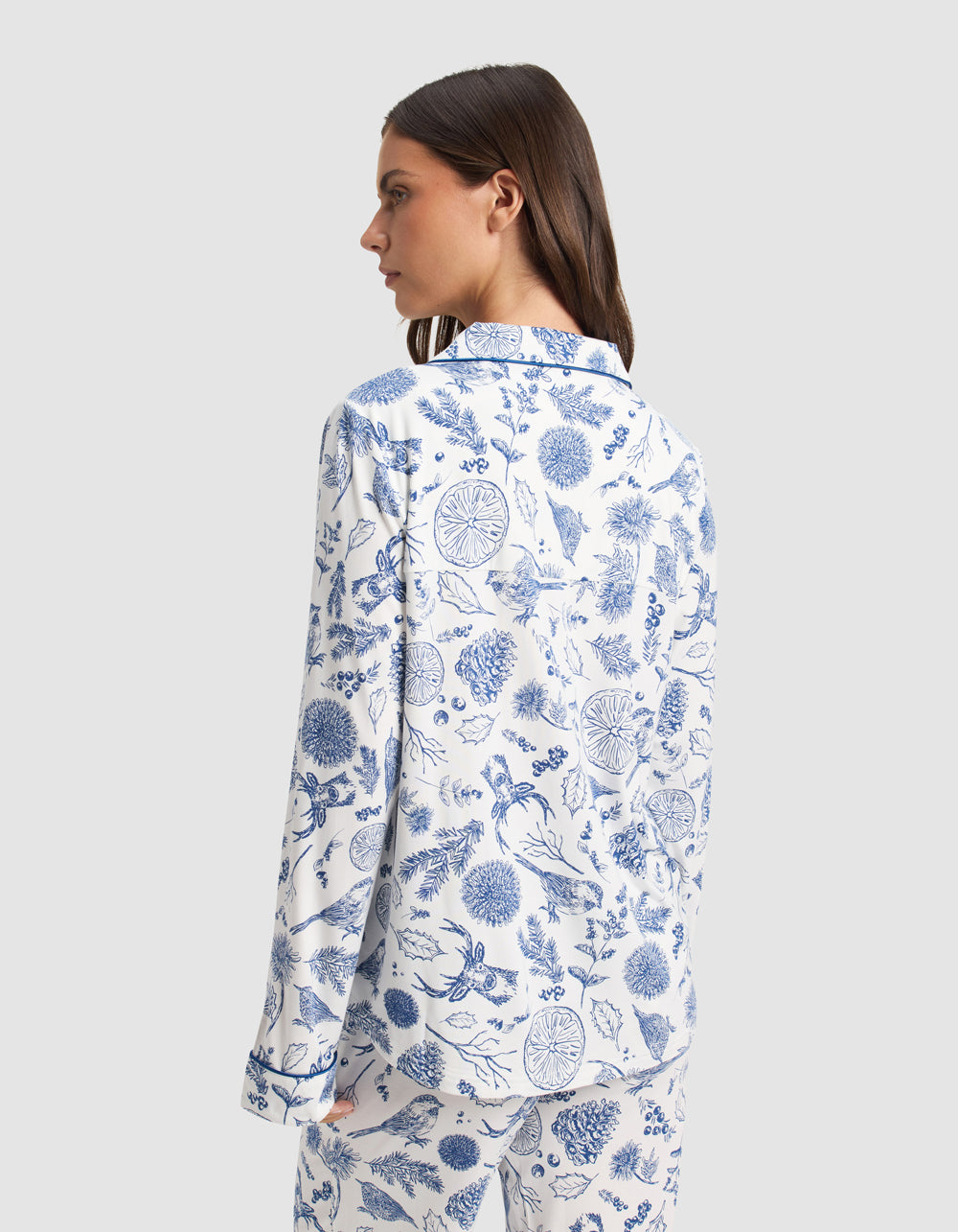 A woman with straight brown hair models the Cozy Earth Women's Bamboo Stretch-Knit Long Sleeve Pajama Top in white with a blue botanical print, standing with her back to the camera against a plain light gray background.