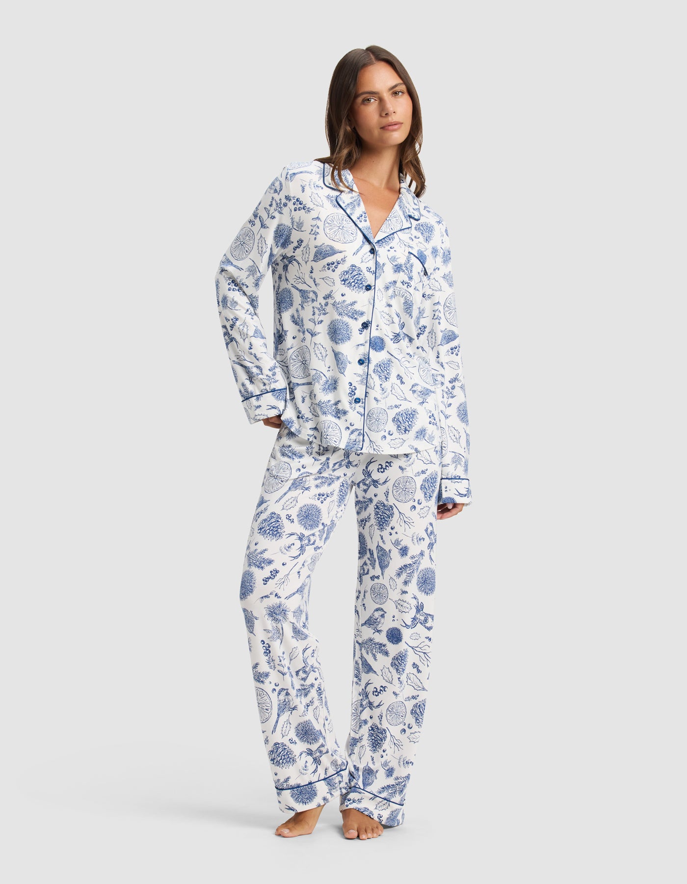 A woman poses barefoot against a light gray background, wearing Cozy Earth's Women's Bamboo Stretch-Knit Long Sleeve Pajama Top—a white base with blue floral print, paired with matching long pants.