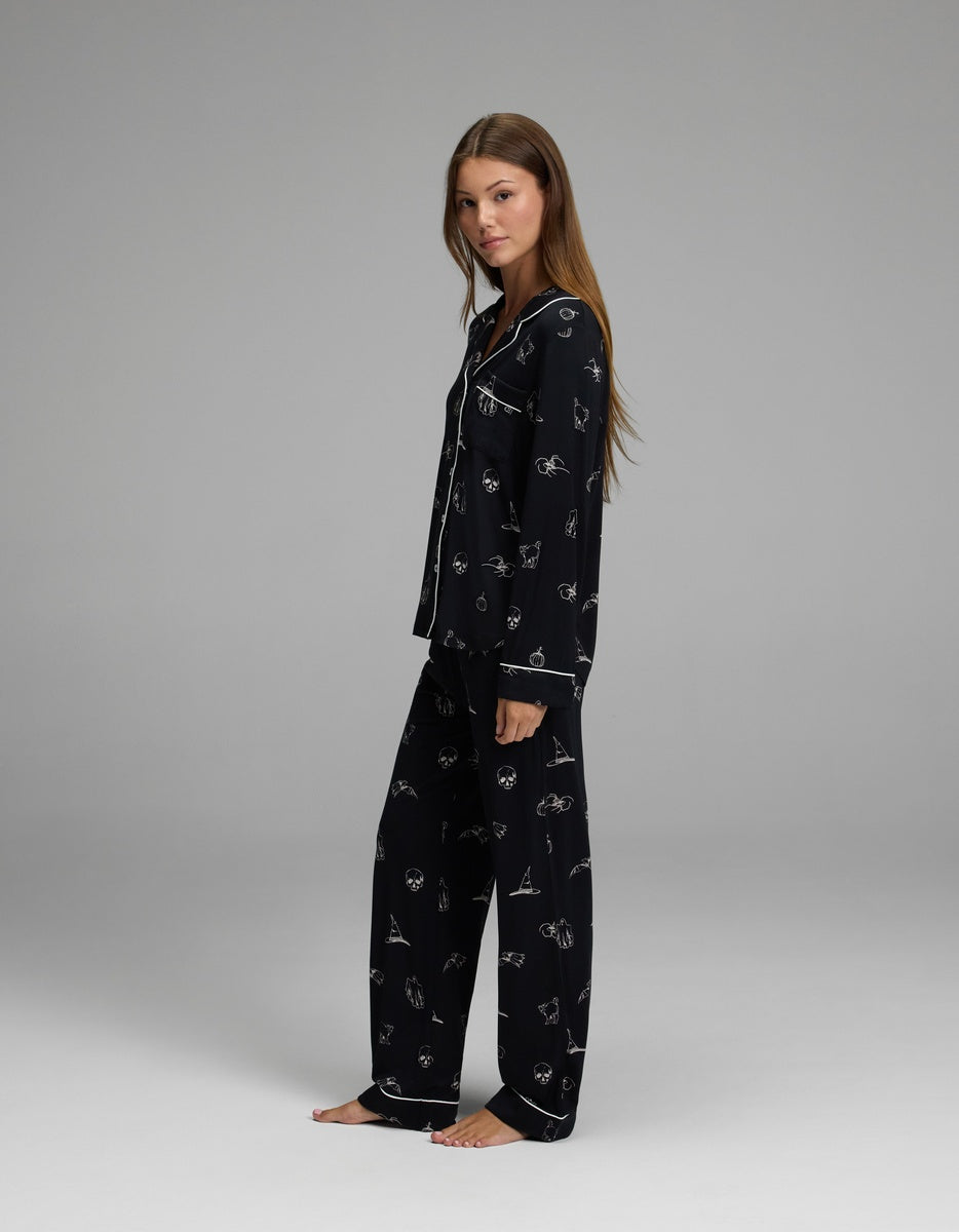 A woman with long brown hair stands barefoot against a plain gray background, wearing the Cozy Earth Women’s Bamboo Stretch-Knit Long Sleeve Pajama Set, featuring matching black pajama pants and a long-sleeved top adorned with white line-drawn designs, as she looks at the camera.