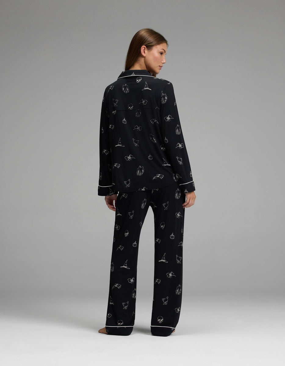 A woman stands barefoot on a gray background, wearing the Cozy Earth Women’s Bamboo Stretch-Knit Long Sleeve Pajama Set in black with a white outline pattern. She faces away but glances over her shoulder, her long hair draped down her back.