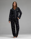 A woman stands barefoot in a relaxed pose, wearing the Cozy Earth Women’s Bamboo Stretch-Knit Long Sleeve Pajama Set—black with white piping and small white illustrated patterns—against a plain gray background.|Color:Halloween Print