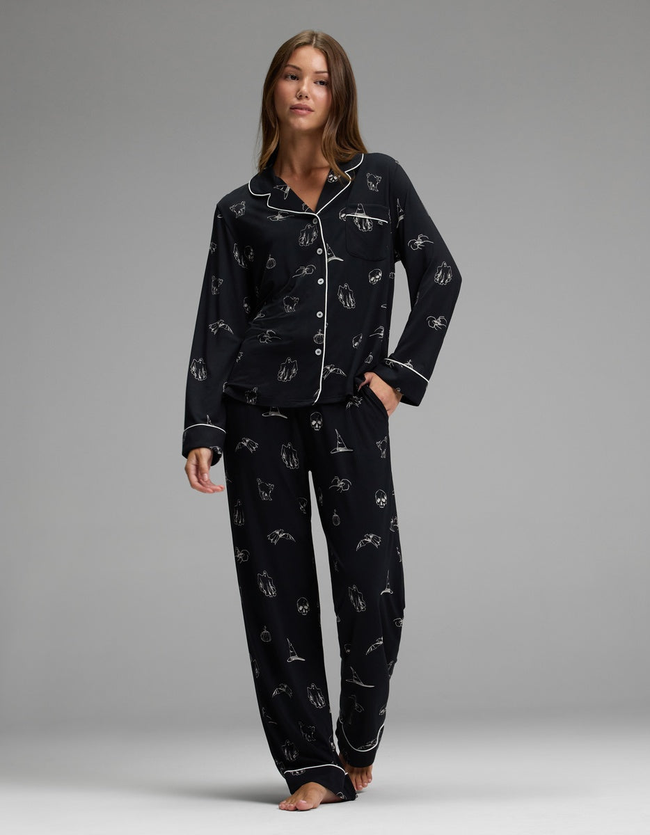A woman stands barefoot in a relaxed pose, wearing the Cozy Earth Women’s Bamboo Stretch-Knit Long Sleeve Pajama Set—black with white piping and small white illustrated patterns—against a plain gray background.