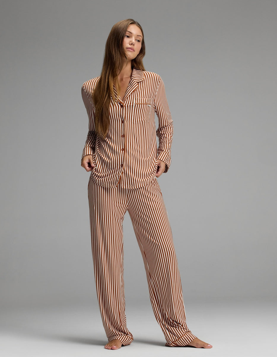 A person stands barefoot on a gray floor, wearing the Cozy Earth Women’s Bamboo Stretch-Knit Long Sleeve Pajama Set in brown-and-white vertical stripes, featuring loose-fitting long sleeves and pants, against a plain gray background.