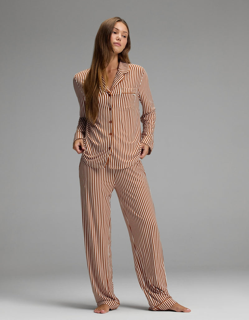 A person stands barefoot on a gray floor, wearing the Cozy Earth Women’s Bamboo Stretch-Knit Long Sleeve Pajama Set in brown-and-white vertical stripes, featuring loose-fitting long sleeves and pants, against a plain gray background.
