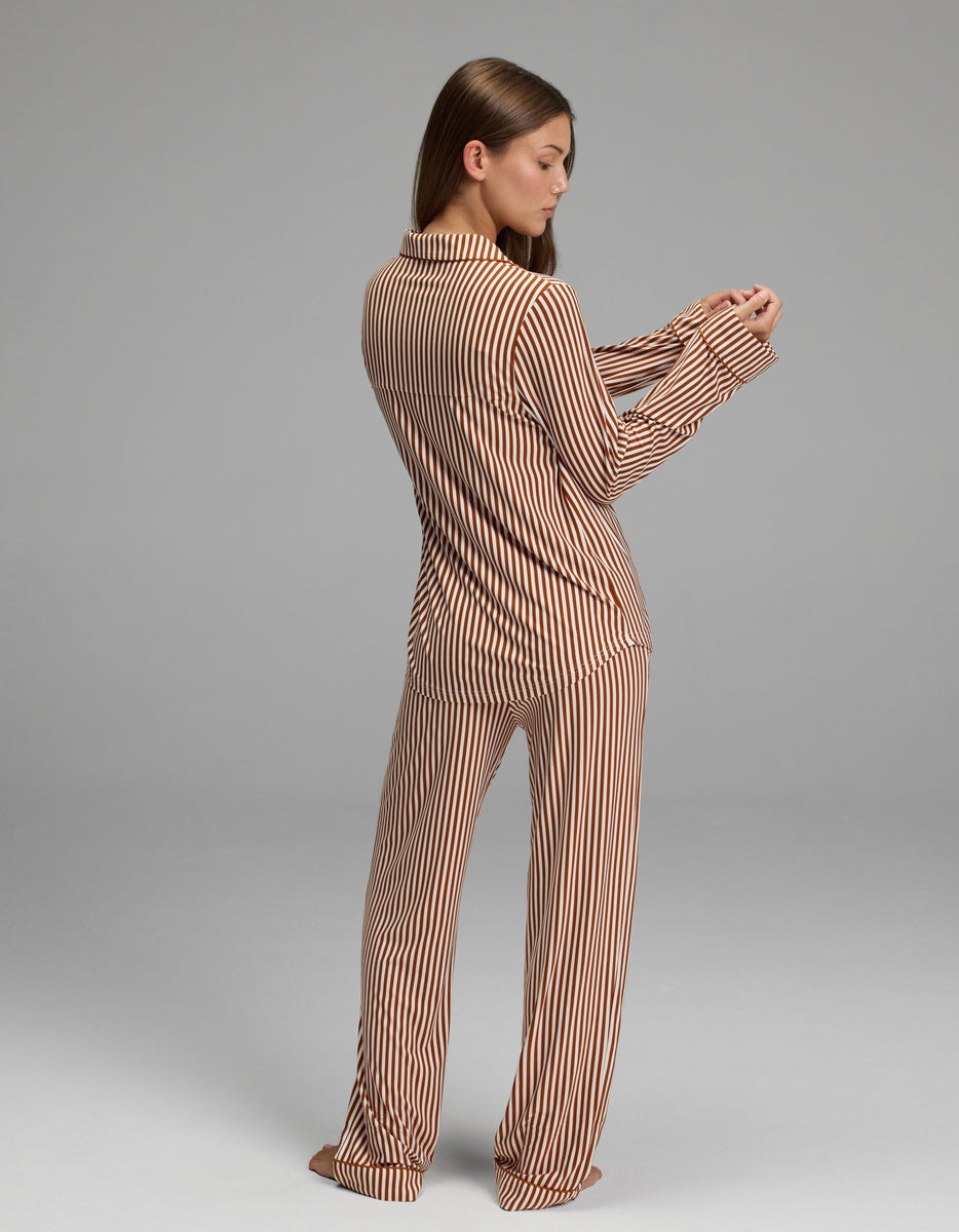 A woman stands barefoot, facing away and looking at her sleeve, wearing the Cozy Earth Women’s Bamboo Stretch-Knit Long Sleeve Pajama Set featuring vertical brown and beige stripes. The background is plain gray.